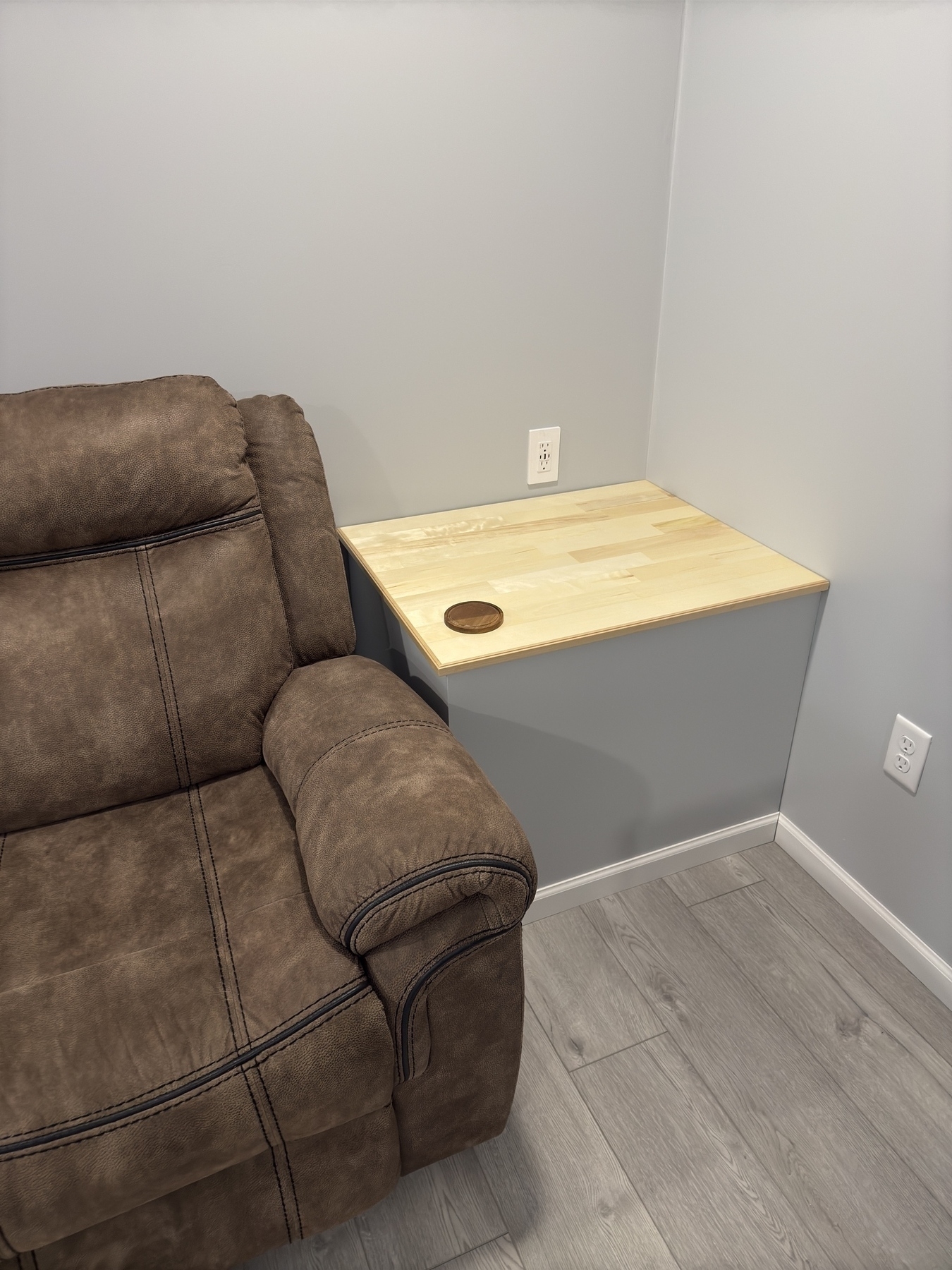 A brown cushioned chair is positioned next to a small wooden table in the corner of a room with light gray walls and laminate flooring.