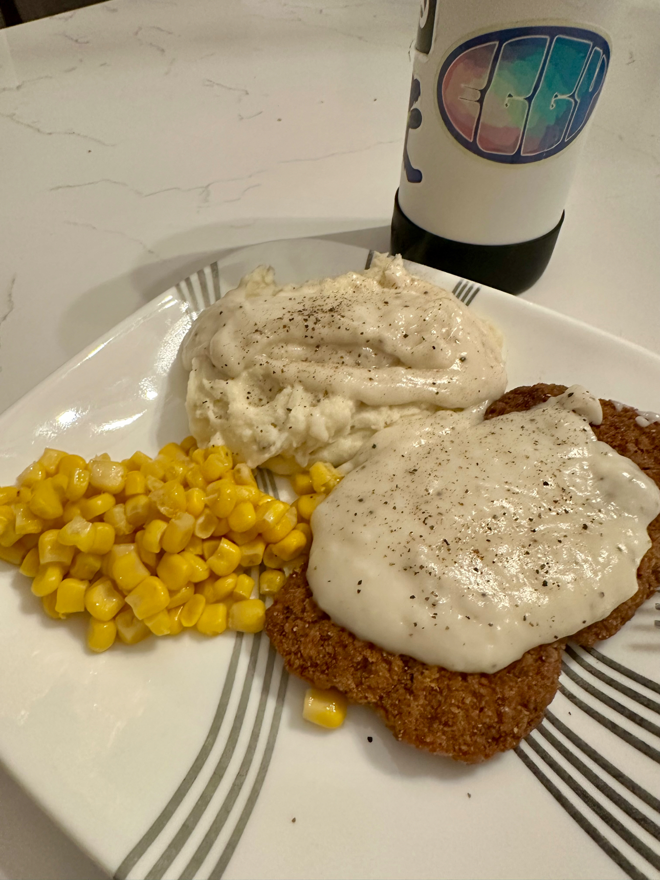 A plate with chicken-fried steak topped with cream gravy, mashed potatoes also with gravy, and a side of corn kernels. In the background, a bottle with a colorful logo.