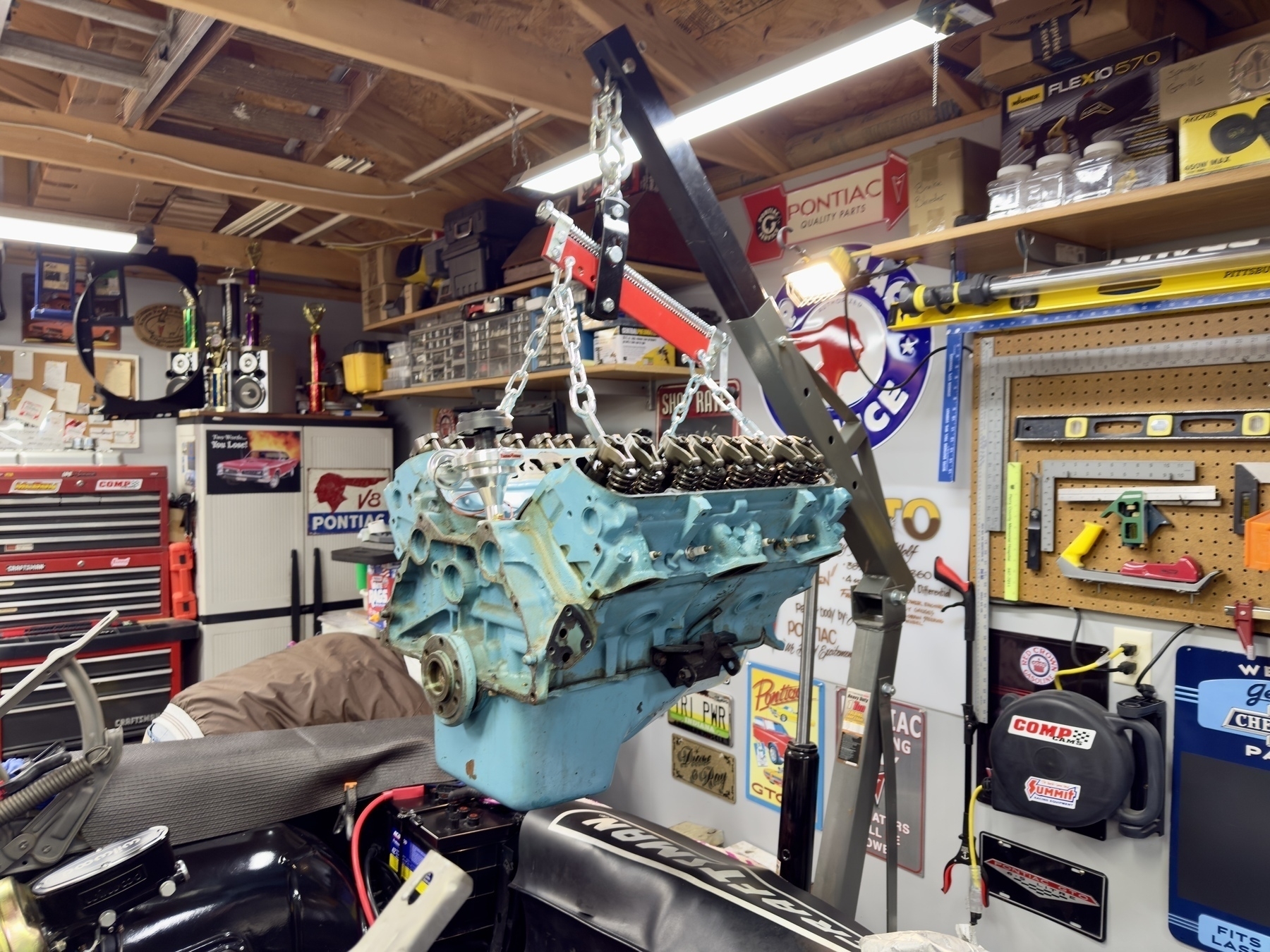 A blue engine block is being hoisted by a crane in a cluttered automotive workshop.