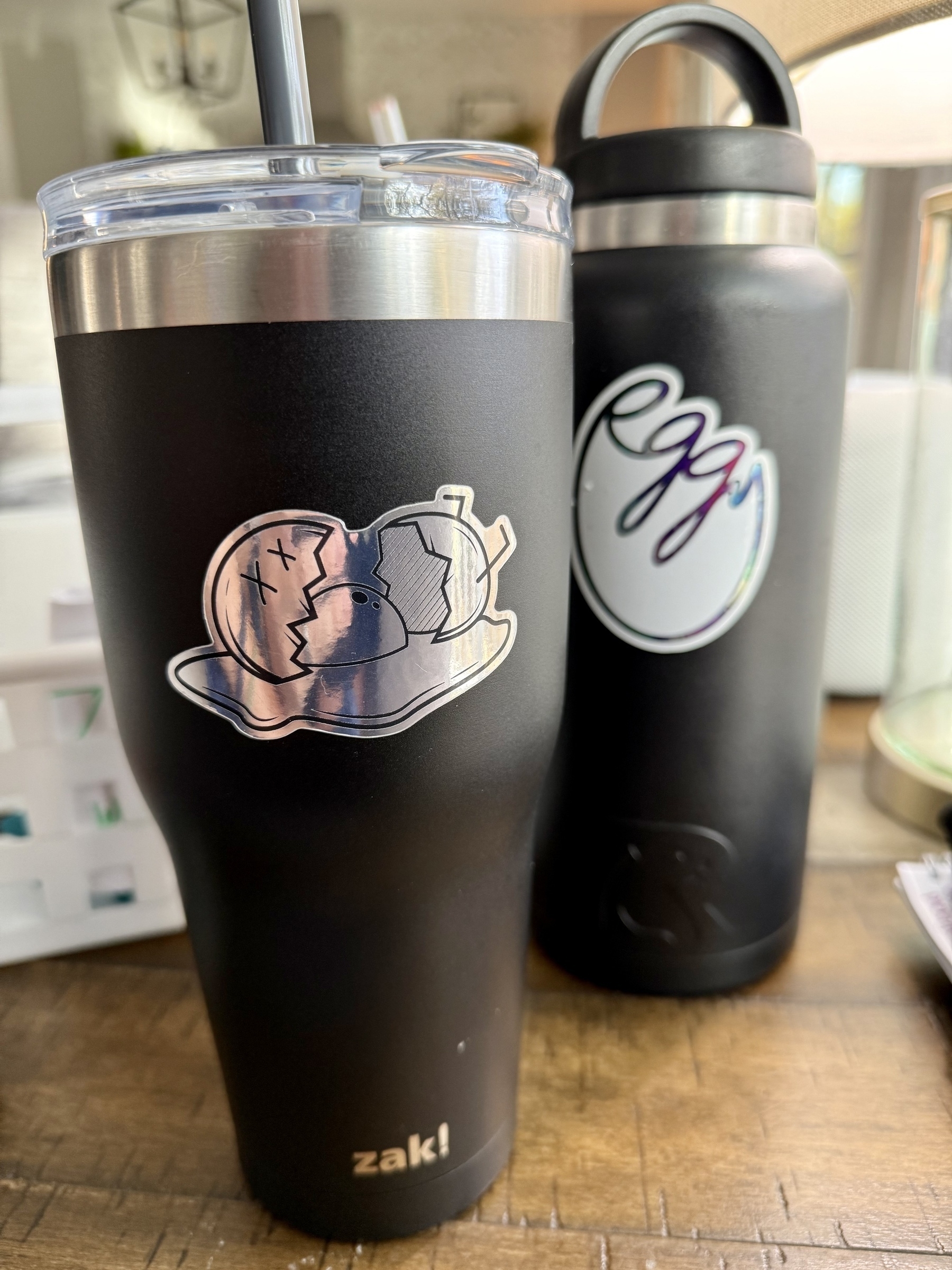 Two black insulated drink containers are displayed, each adorned with unique stickers and a metal rim, set on a wooden surface.