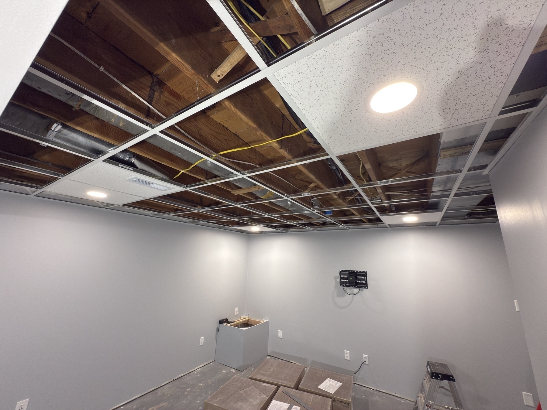 A room under construction features an open ceiling grid with exposed wiring and partial ceiling tiles, along with stacked boxes and wall-mounted equipment.