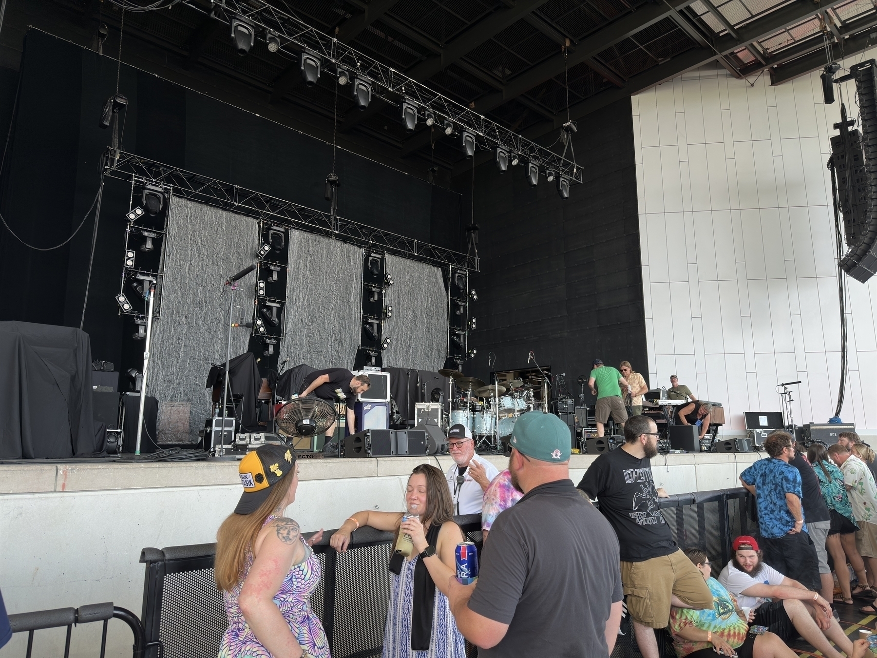 A group of people are gathered in front of an outdoor stage with musical equipment set up, preparing for a performance.