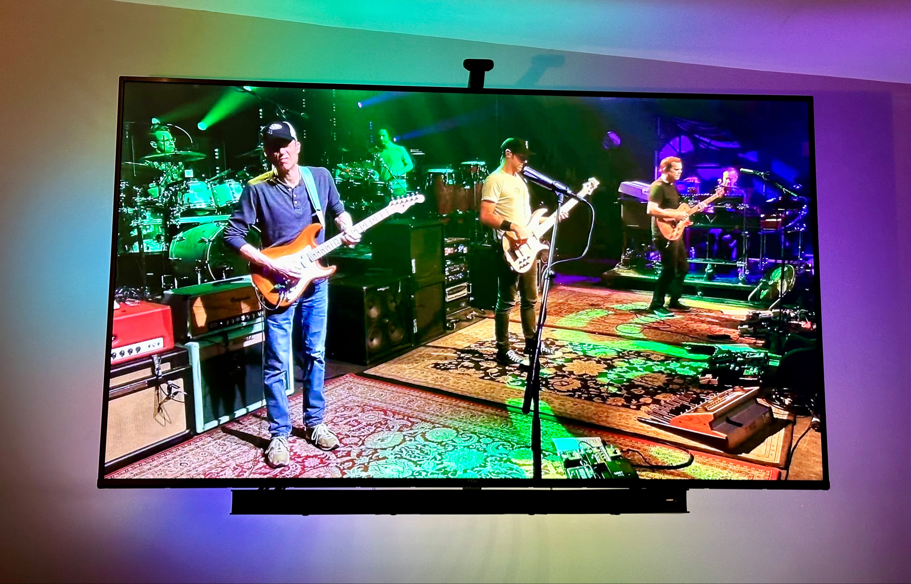 A television screen mounted on a wall displaying a live concert with musicians playing guitars and a drummer in the background.