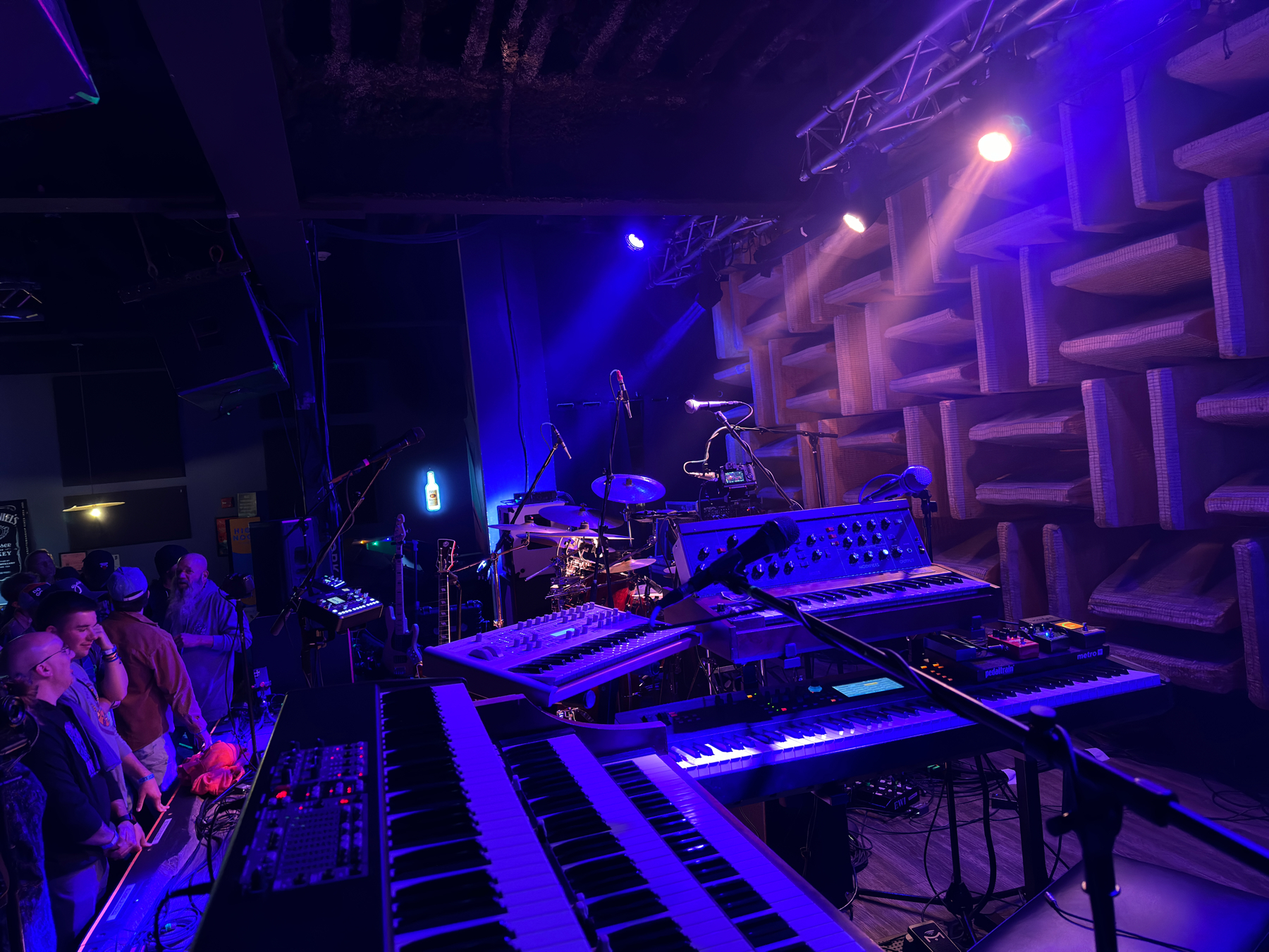 A music venue stage with various electronic keyboards and synthesizers set up for a performance. Audience members gather in anticipation. The stage is illuminated with blue stage lighting and features acoustic panels on the wall for sound dampening.