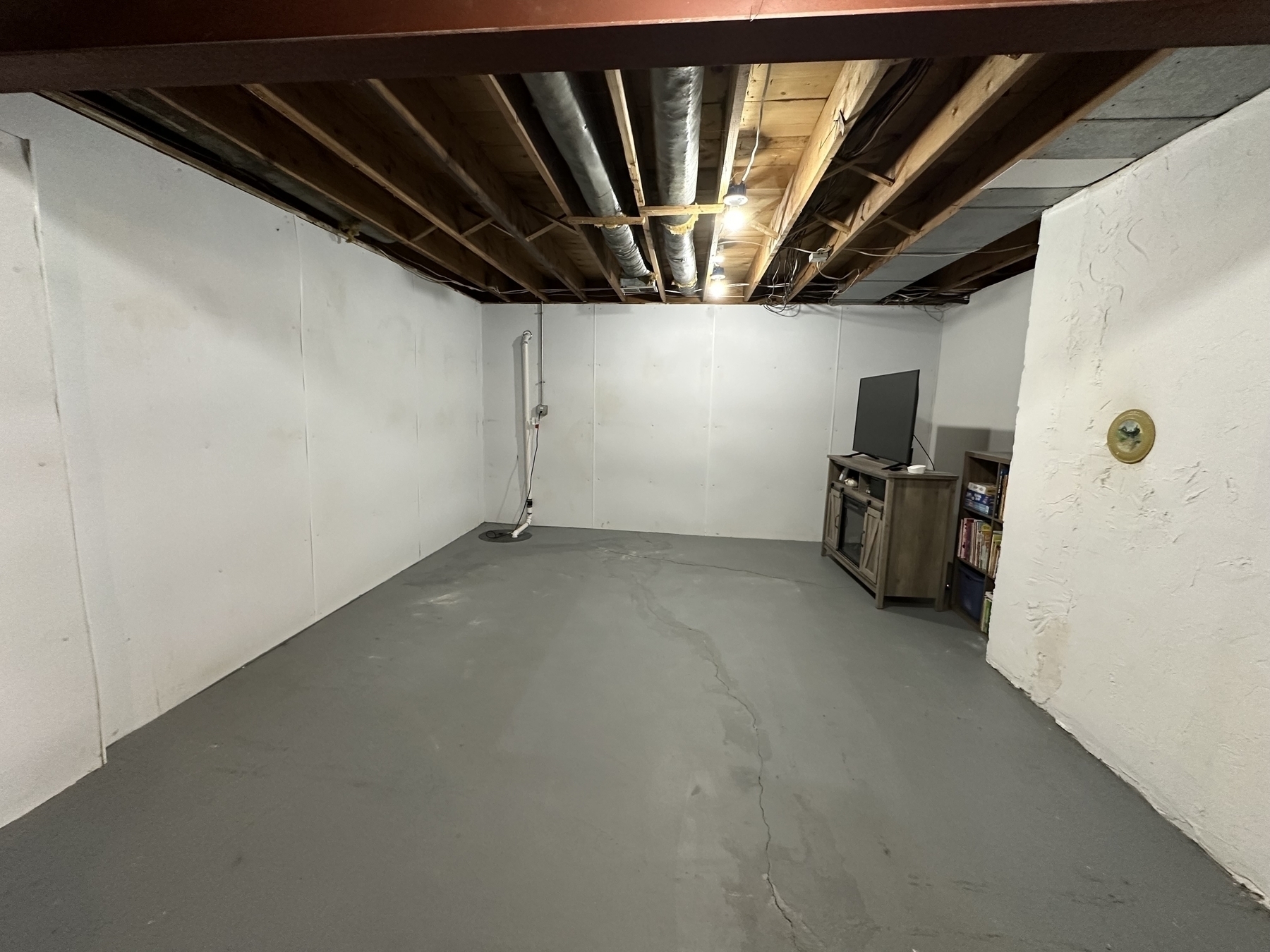 Auto-generated description: A basement room features exposed ceiling beams, a concrete floor, and a corner entertainment setup with a television on a stand.