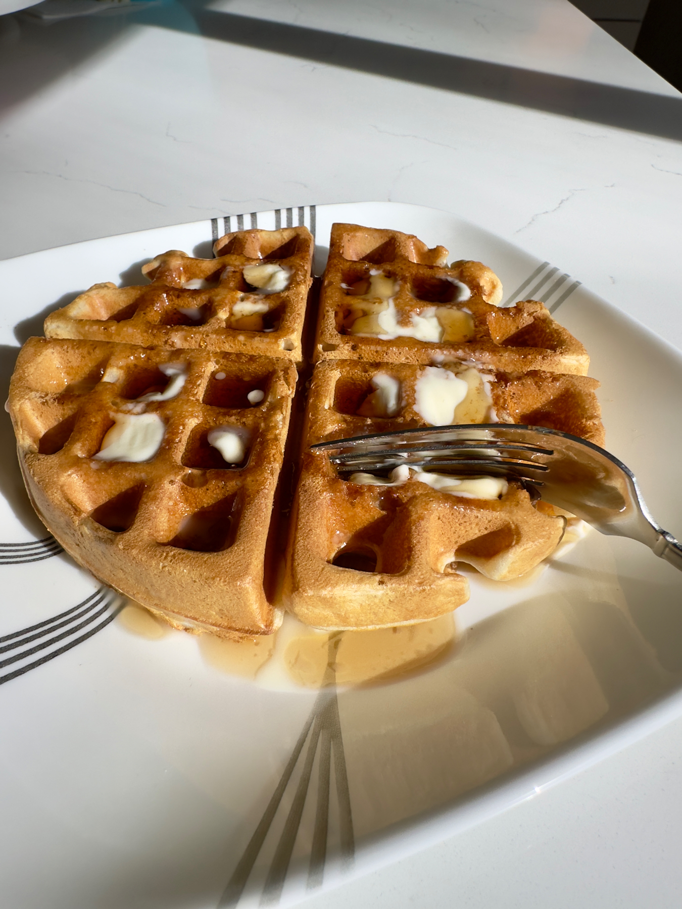 A waffle cut into quarters on a white plate, drizzled with syrup and butter, with a fork on the side.