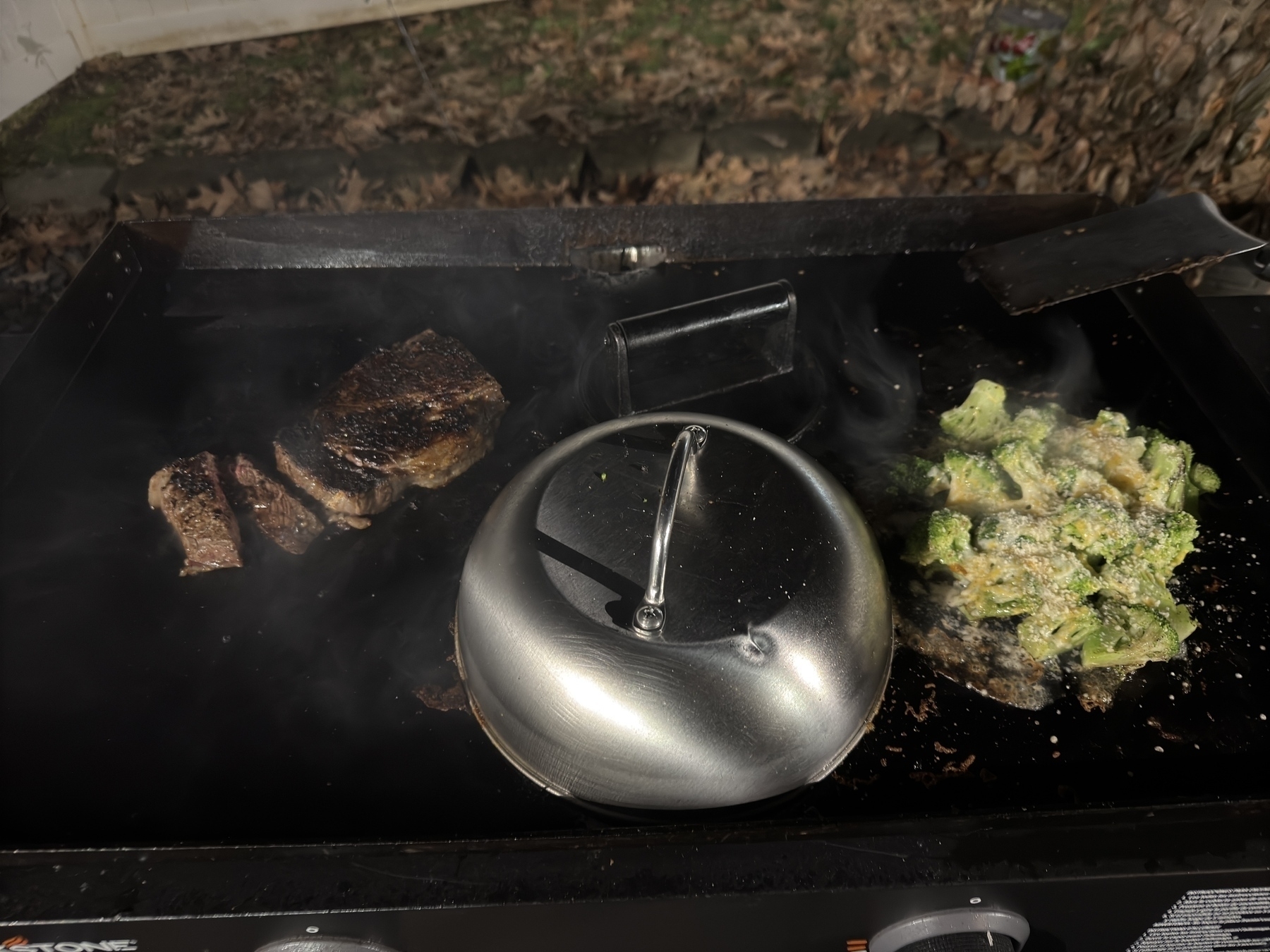 A grill with sizzling steak slices and broccoli under a lid, set against an outdoor background.