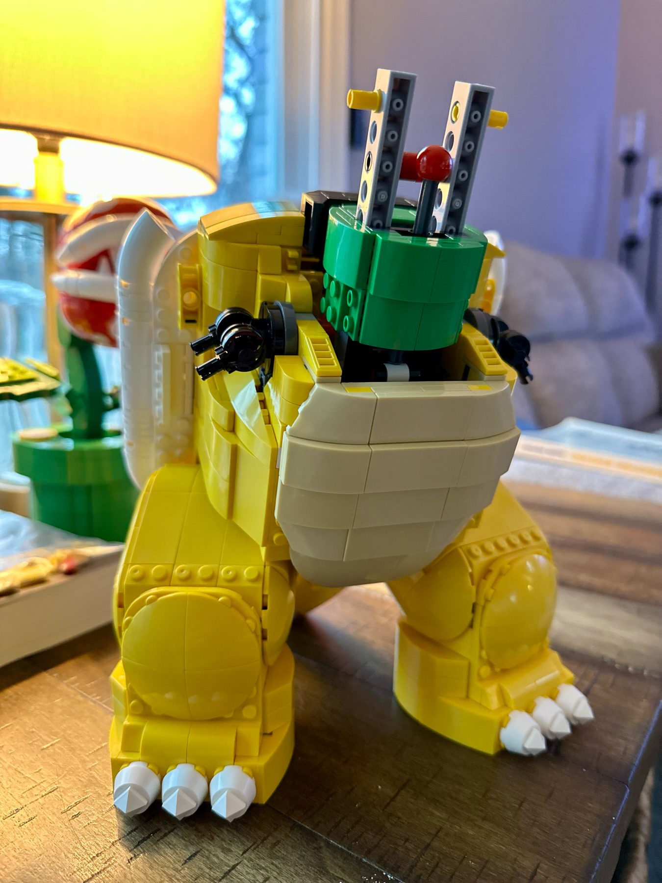 A LEGO model of a dinosaur-like creature with a green and yellow color scheme, featuring a round body, short stubby arms, and a cockpit containing two control handles and a red button. The creature stands on a wooden surface with part of a home