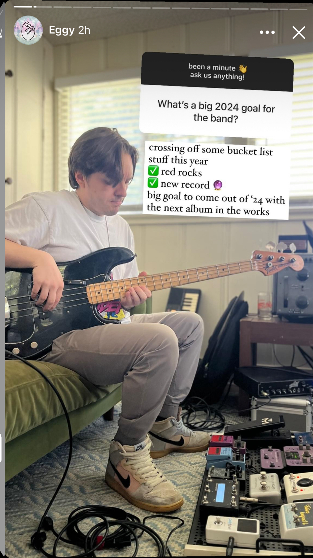 A person seated while playing a black bass guitar, with a variety of guitar pedals at their feet. The individual is focused on the instrument, surrounded by music equipment. A text overlay reveals goals for a band in 2024, including playing at Red