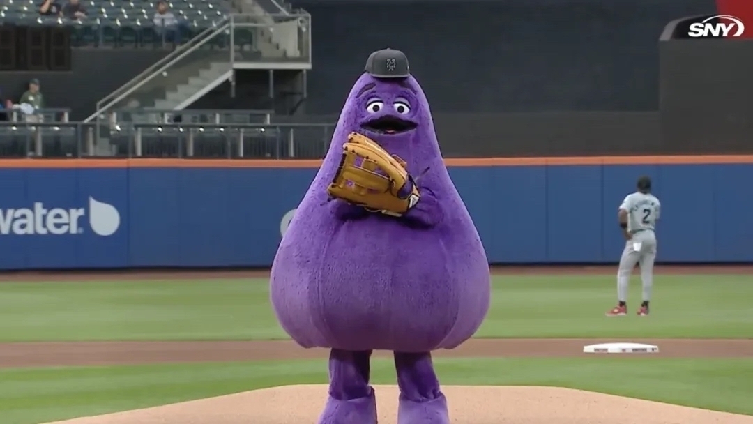 A large, purple mascot is standing on a baseball pitcher's mound holding a baseball glove.