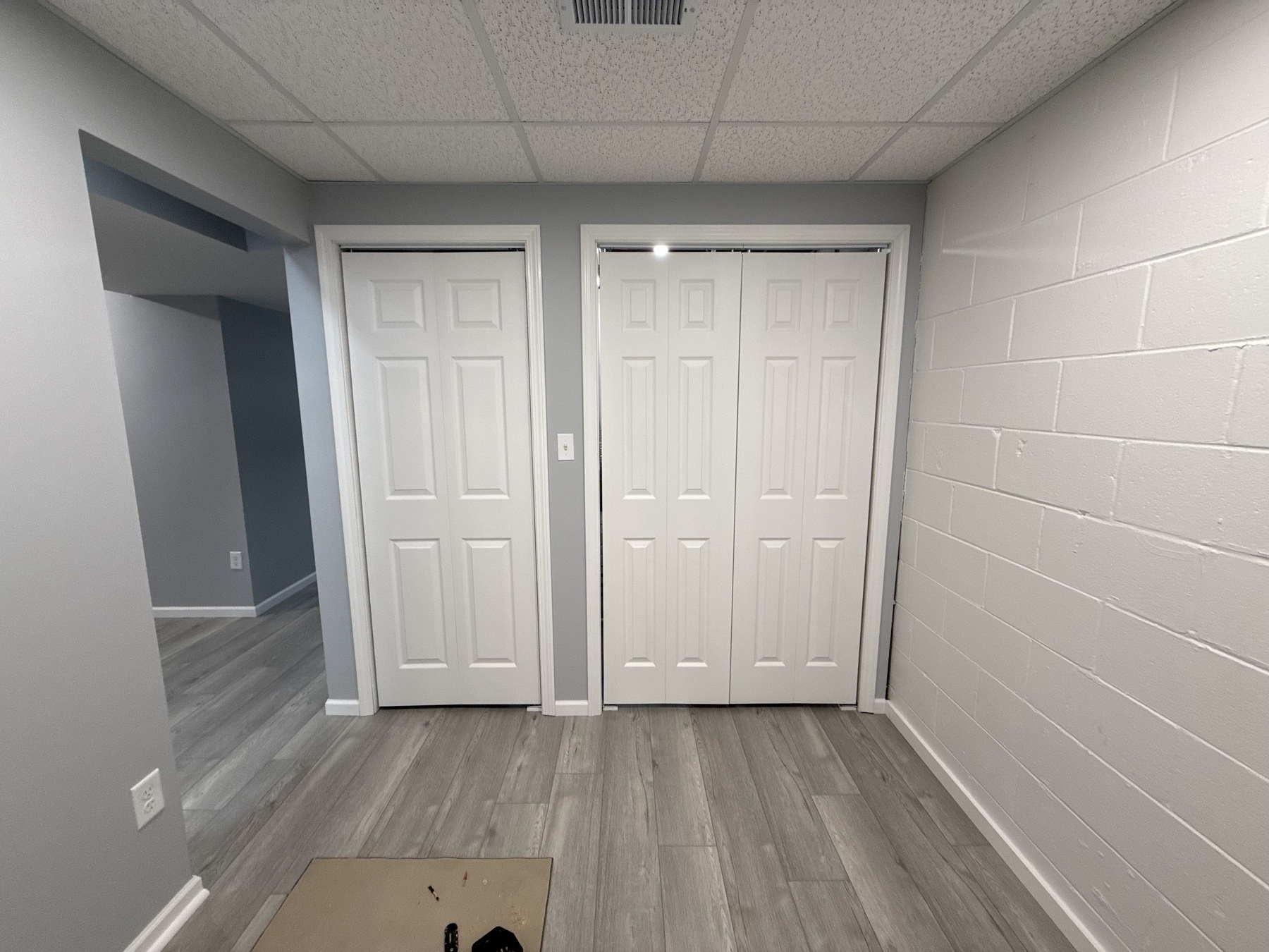 A finished basement area features two closed white closet doors, a partially visible hallway, and gray-toned flooring with white brick wall accents.