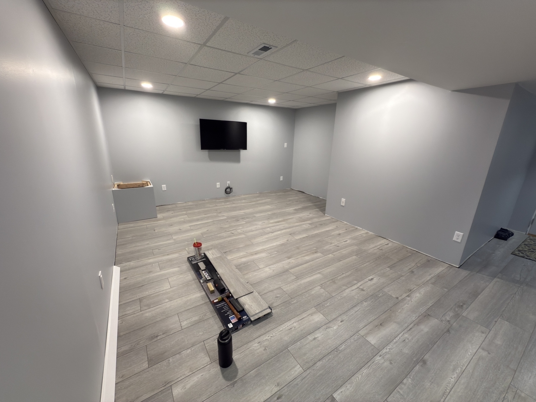 A grey-painted room features a wall-mounted TV, recessed lighting, and light wood flooring, with a few items placed on the floor.