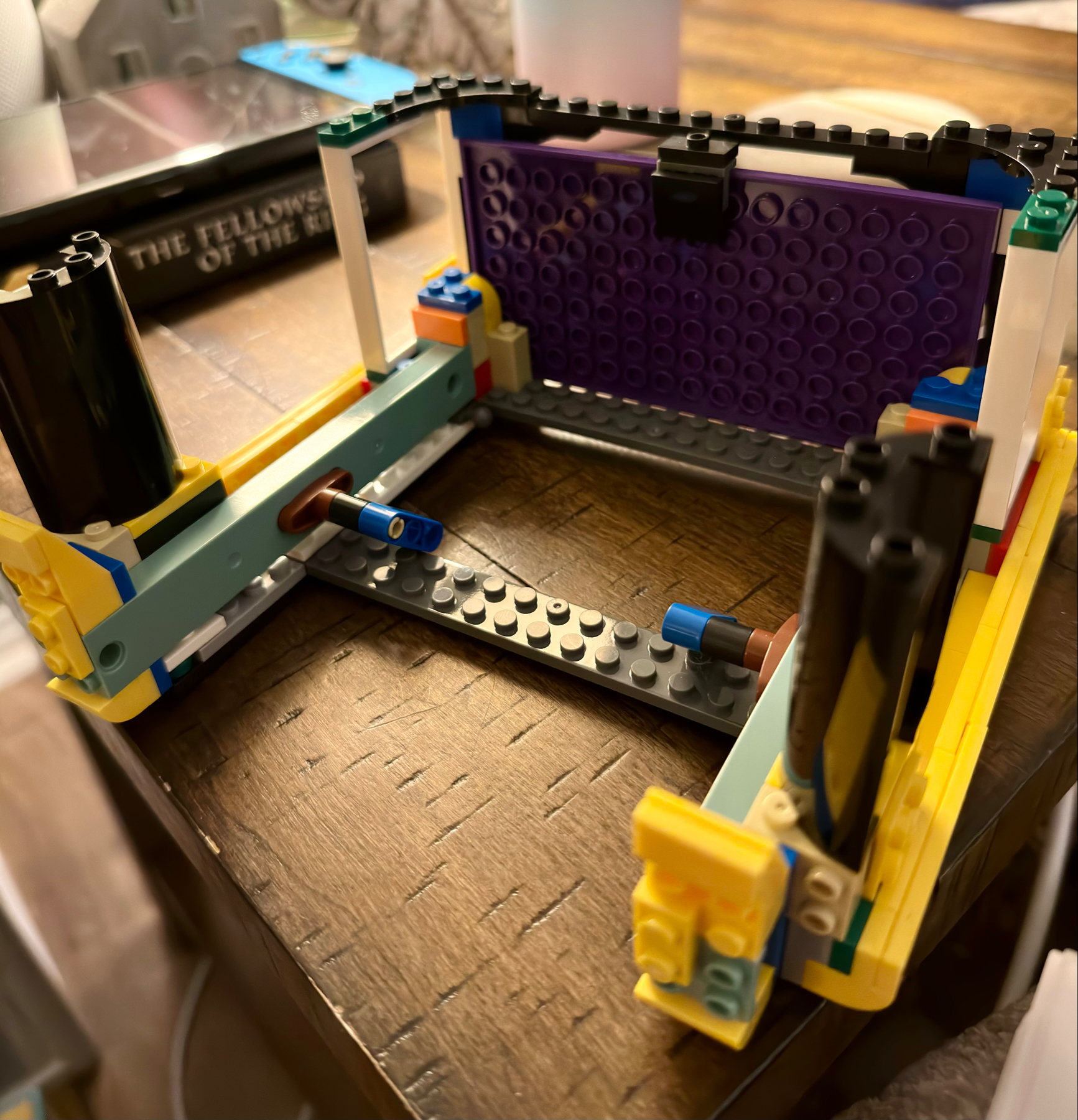 A partially assembled LEGO structure showcasing various colorful pieces and bricks on a wooden surface. The construction features black, yellow, blue, green, and purple elements, indicating an early stage of the building process.