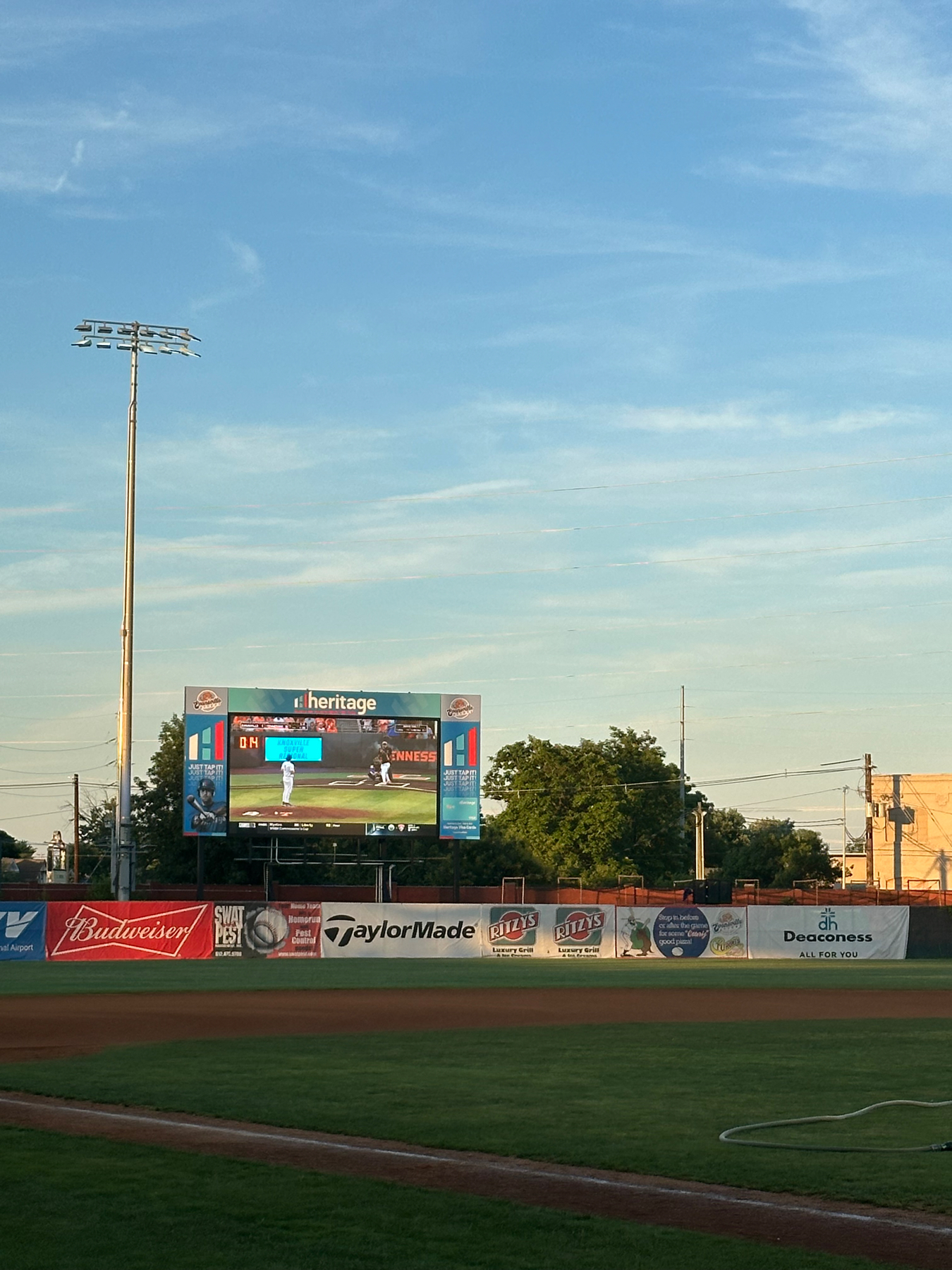 A baseball stadium with a large digital scoreboard displaying game information and advertisements. The field is green with a well-maintained infield and outfield. Advertising banners are visible along the outfield fence. The sky is clear with light clouds, and a tall