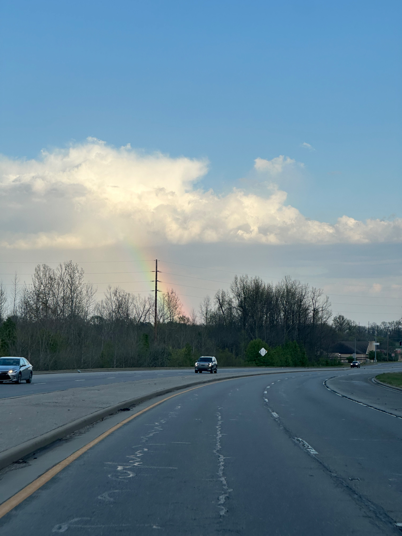 A road curving to the right with vehicles in the distance and a faint rainbow appearing in the partial cloud cover against a blue sky.