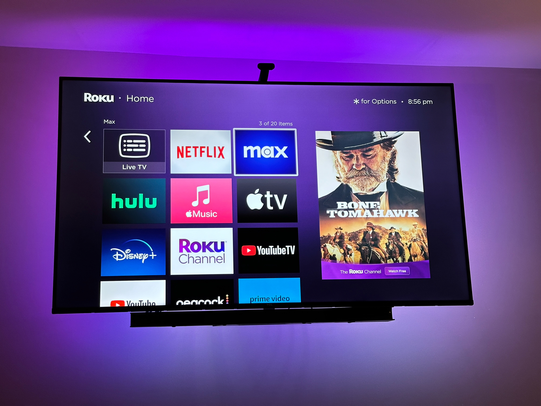 A television mounted on a wall displays the Roku home screen, featuring app icons such as Netflix, Hulu, Disney+, and others. On the right side, there is a highlighted movie poster for “Bone Tomahawk” visible on The Roku Channel section
