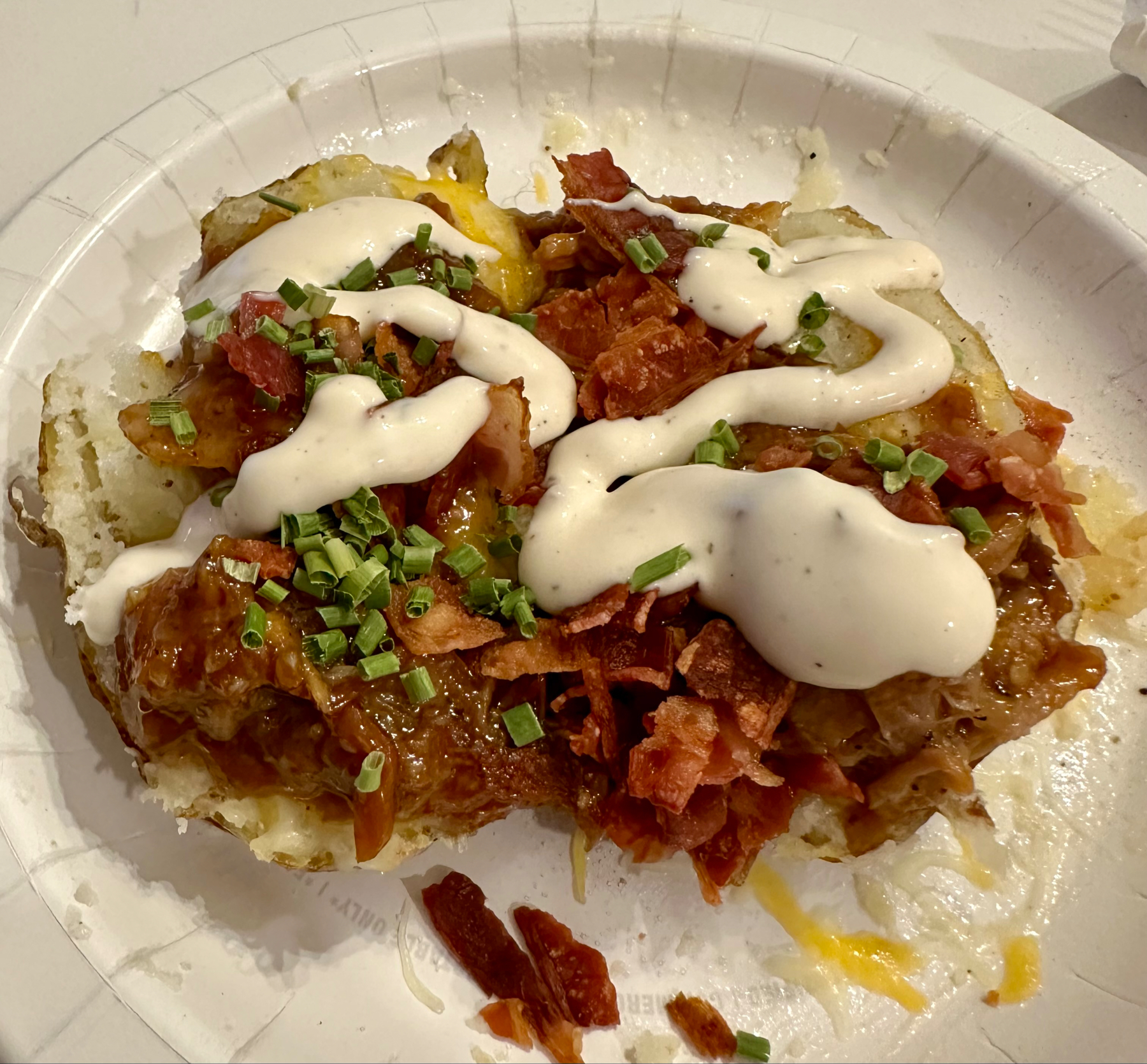 A loaded baked potato with toppings including shredded cheese, bacon bits, chopped green onions, and a drizzle of white sauce on a white disposable plate.