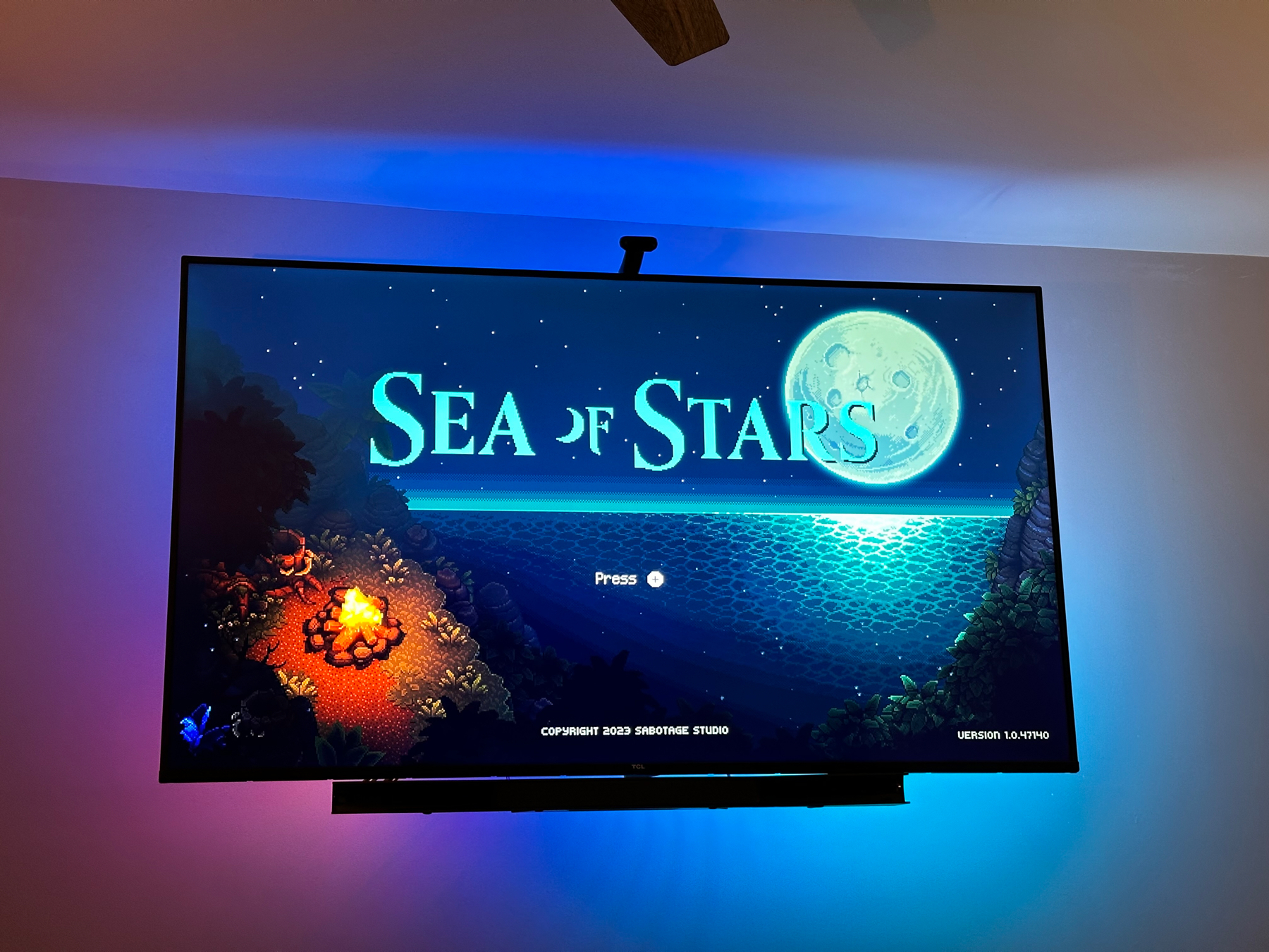 A television mounted on a wall displaying the title screen of the “Sea of Stars” video game, with stylized pixel art graphics showing a moonlit seascape. The room lighting casts a blue ambiance above the TV.