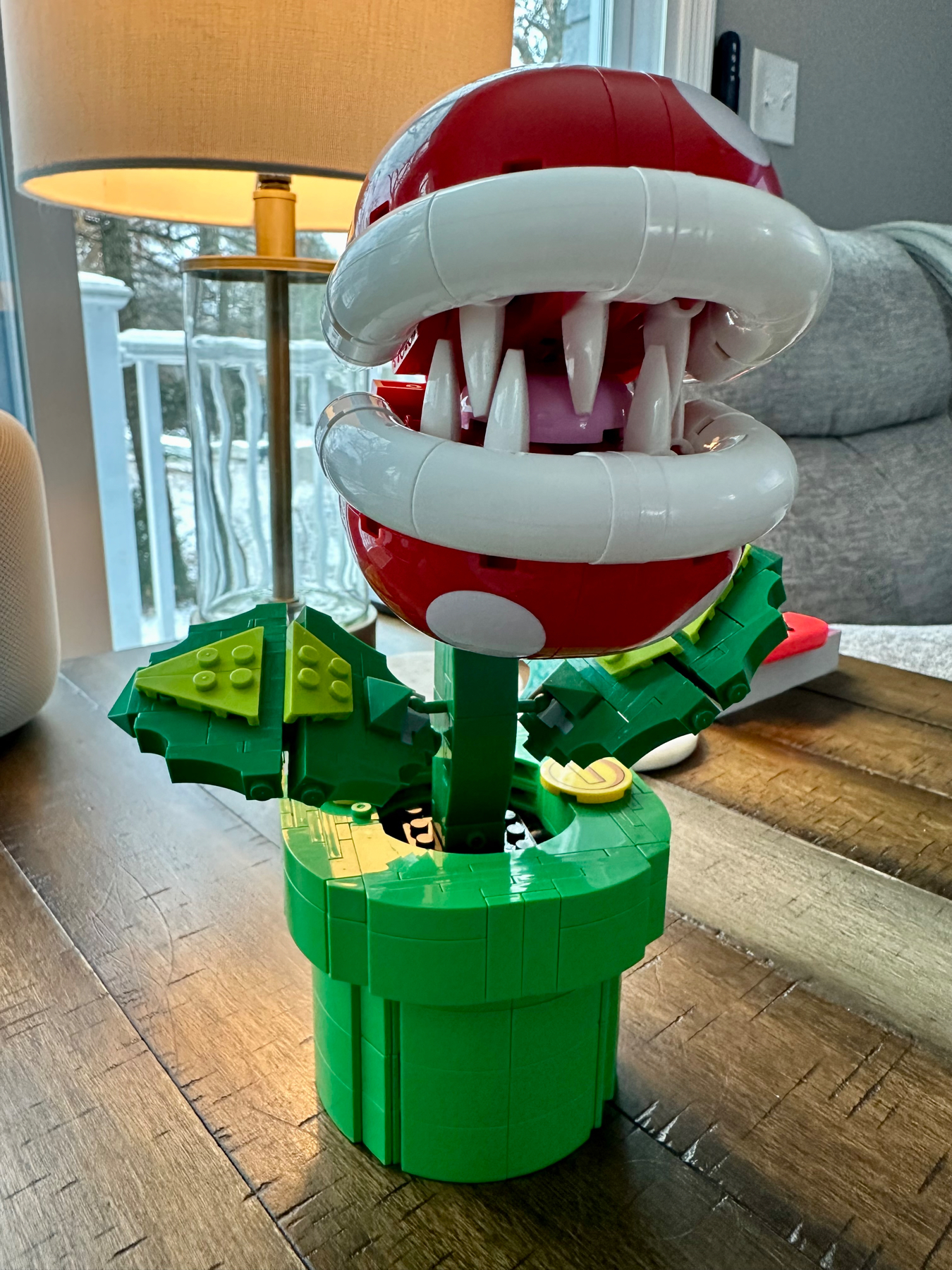 A LEGO set model of a Piranha Plant from the Super Mario series displayed on a wooden floor with a lamp and a window showing a snowy scene in the background.