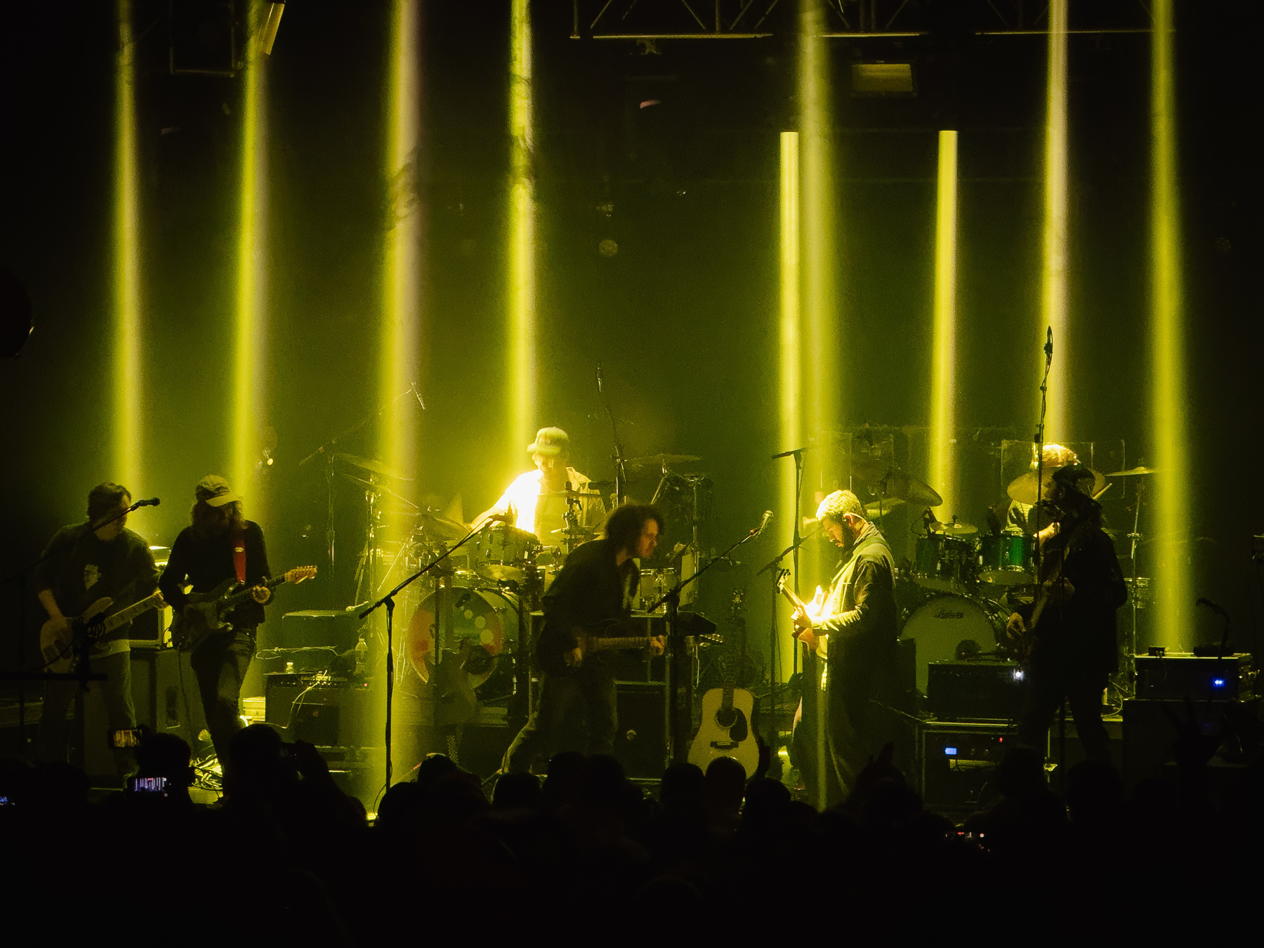 A band is performing on stage with yellow lights illuminating them and musical instruments in the background.