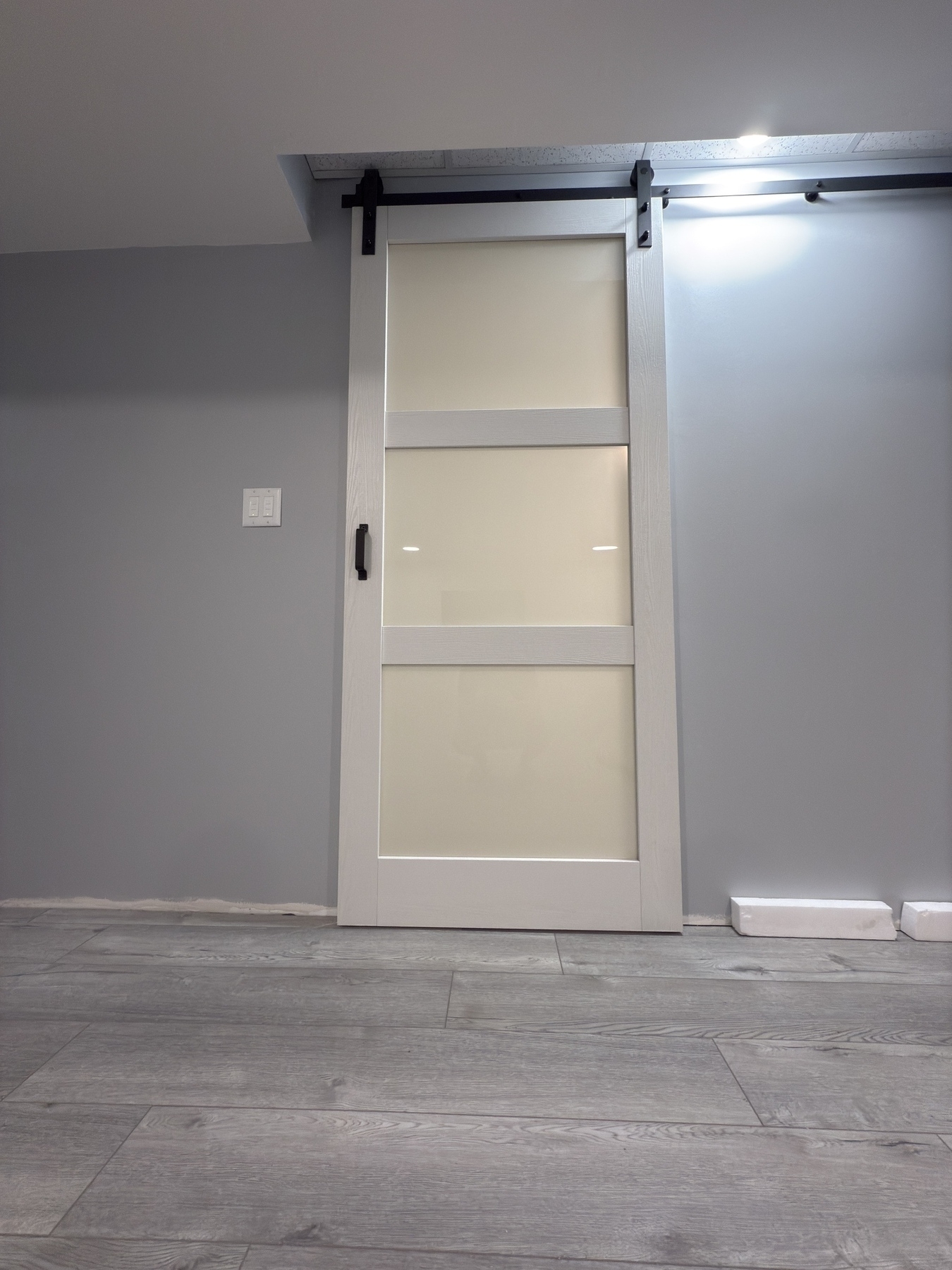 A sliding barn door with frosted glass panels is installed in a modern room with gray flooring and walls.