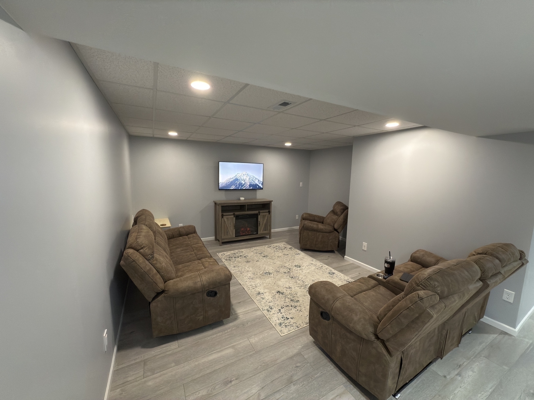 A cozy basement living room with brown reclining sofas facing a wall-mounted TV above a fireplace.