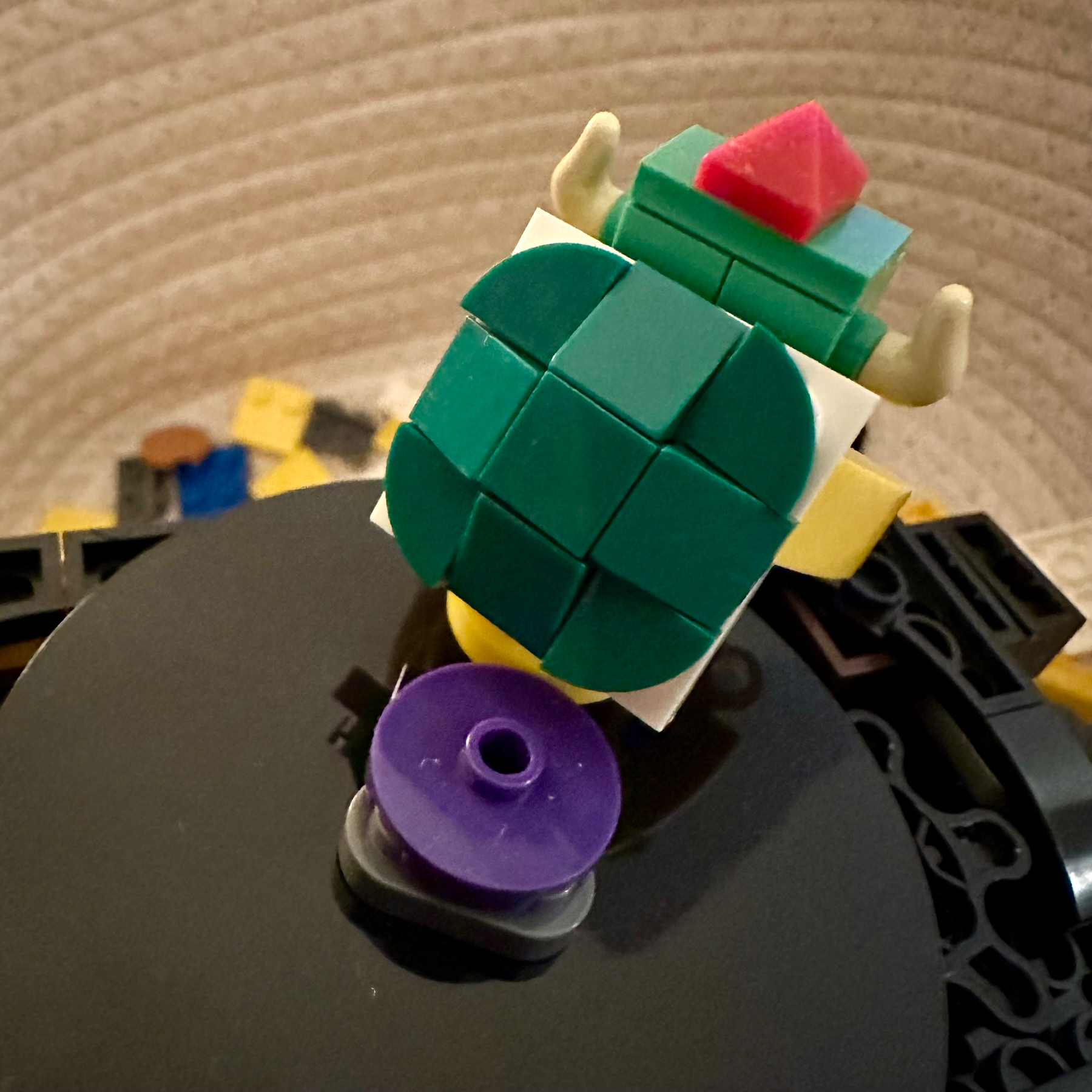 A Lego figure with a green turtle shell backpack and a red accessory on top, positioned on a black and gray base with colorful Lego bricks in the background.