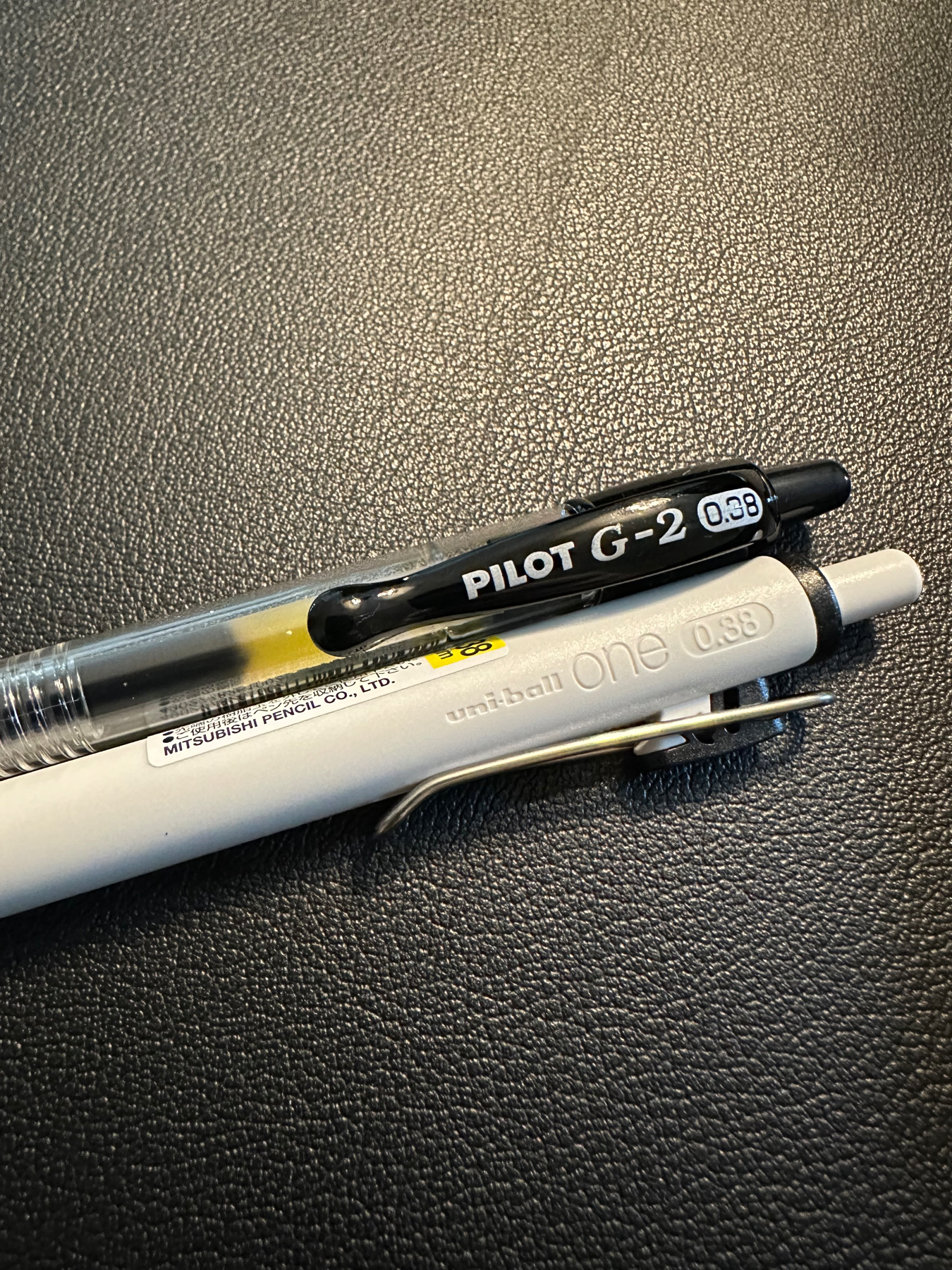 Two pens resting on a textured black surface, the top one is a black Pilot G-2 0.38 and the bottom one is a white Mitsubishi Uniball One 0.38.