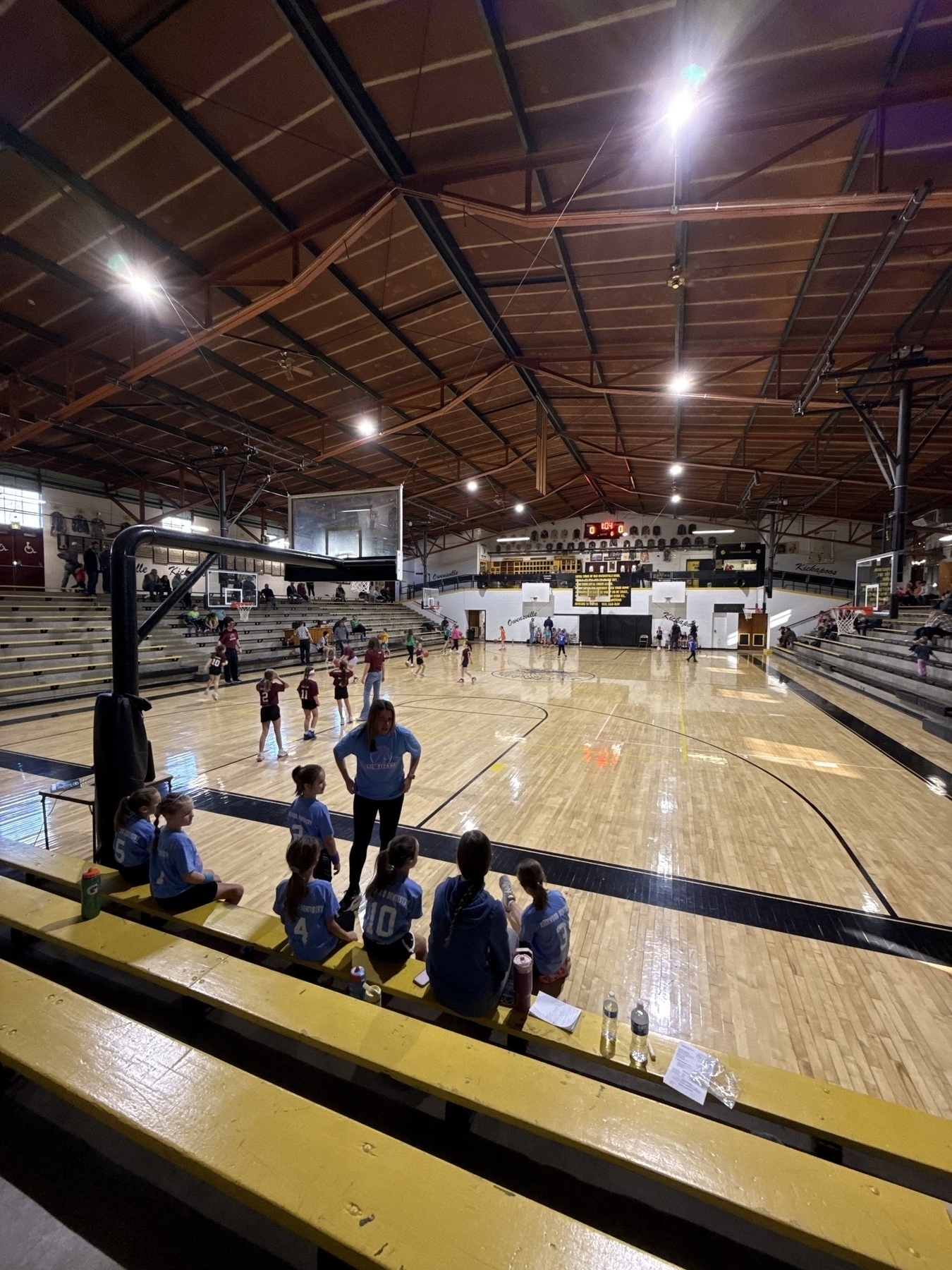 A group of young basketball players sits on a bench inside a gymnasium with multiple games happening on the courts.