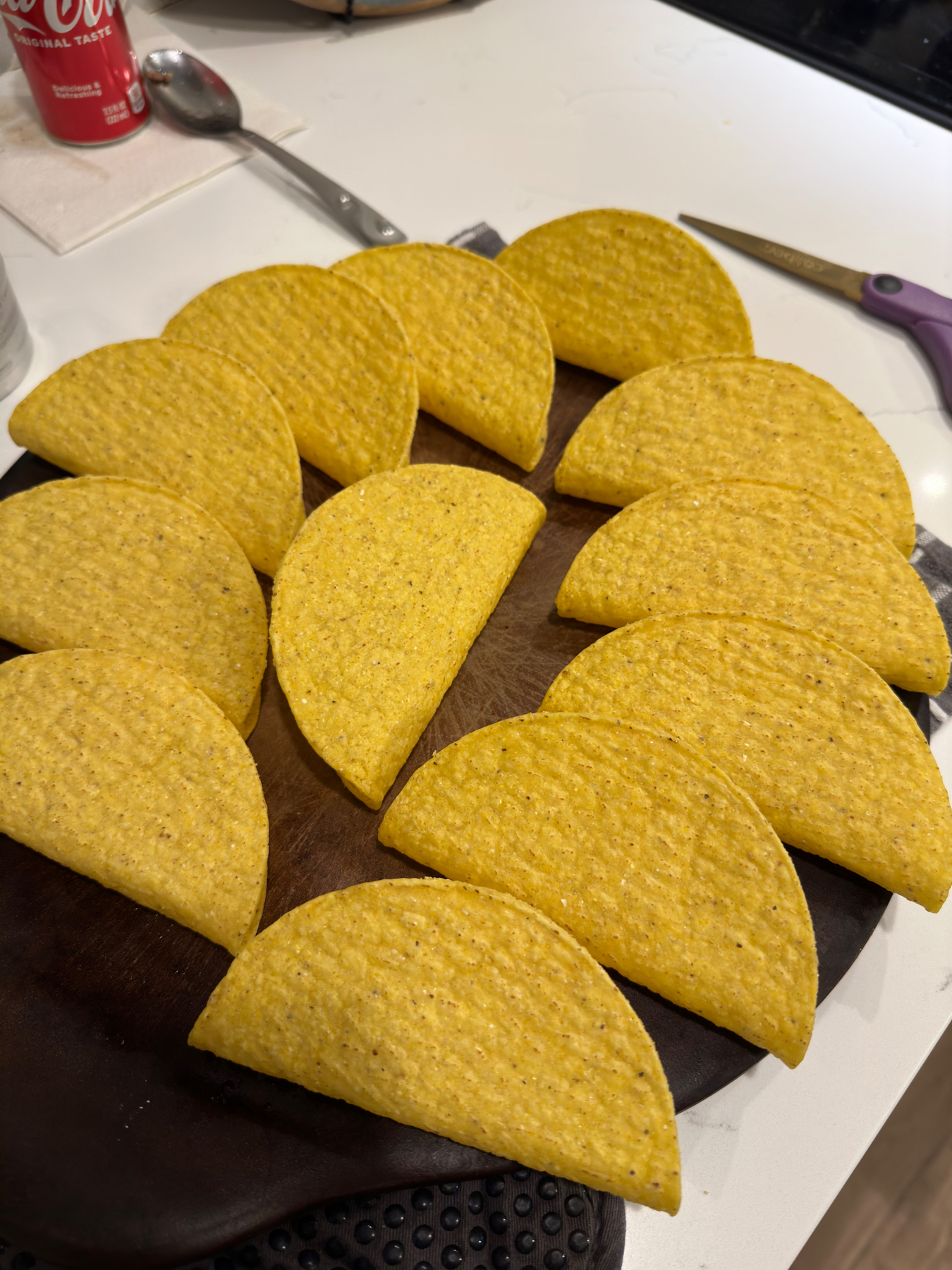 A collection of taco shells spread out on a cutting board with a Coca-Cola can and utensils in the background.