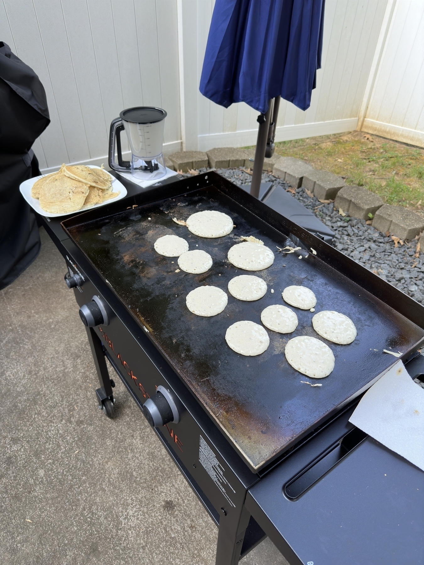 A griddle is being used to cook small, round pancakes next to a plate of prepared pancakes and a kitchen appliance outside.