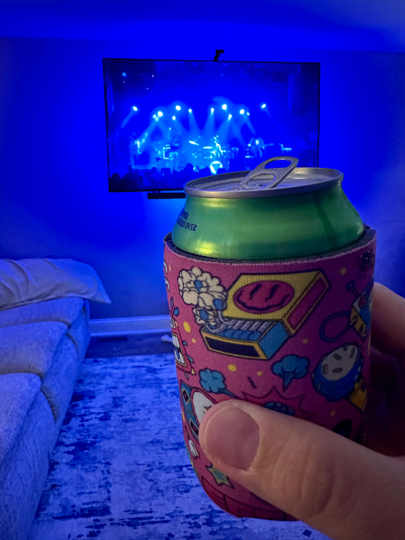 A hand holding a green can with a colorful cozy, in front of a TV displaying a live concert, with LED blue lights illuminating the room.