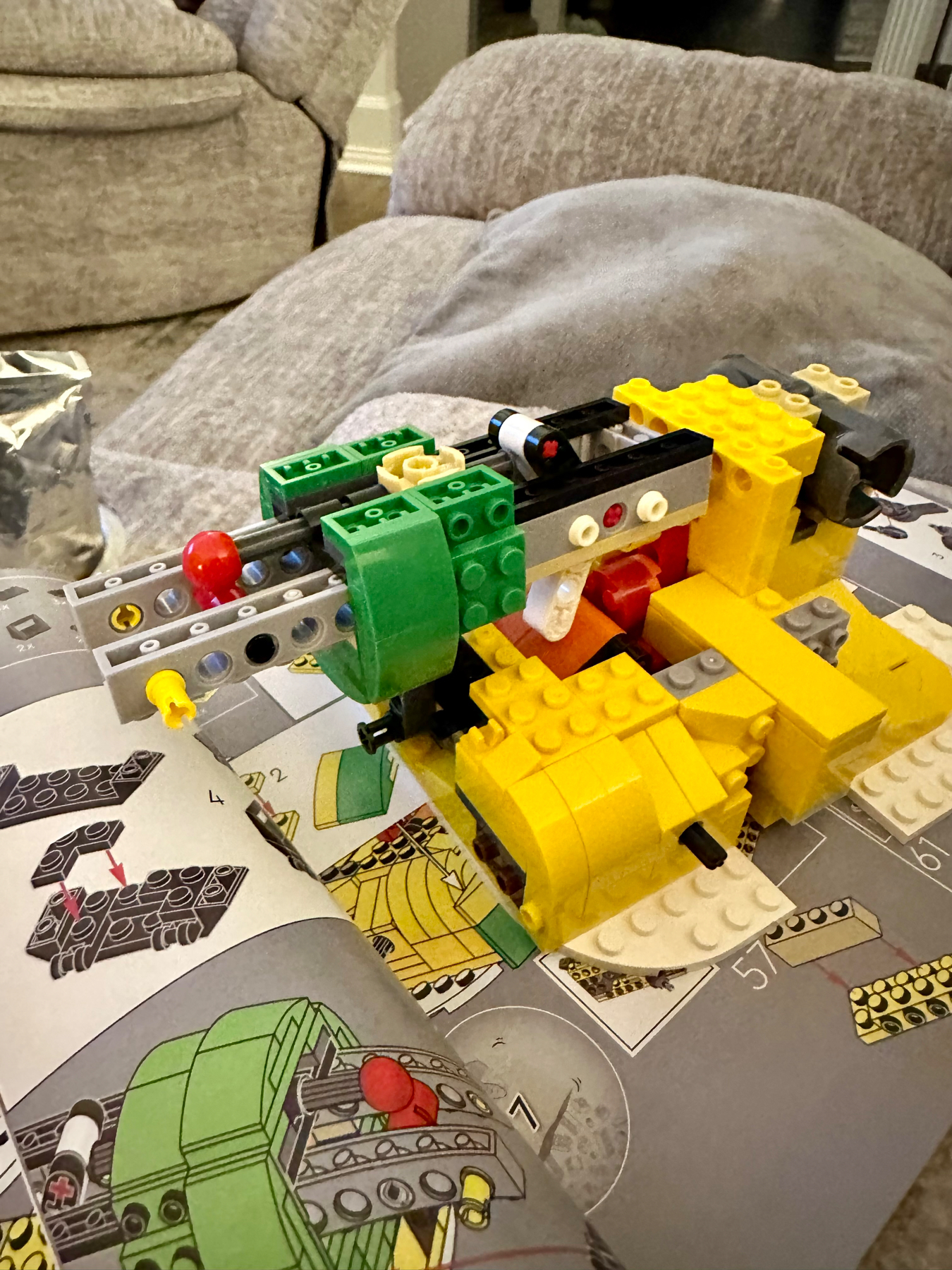 A partially assembled LEGO set resembling a robotic creature or vehicle with green, yellow, and red pieces, resting on an open instruction manual, with a blurred background of a cushioned chair and a blanket.