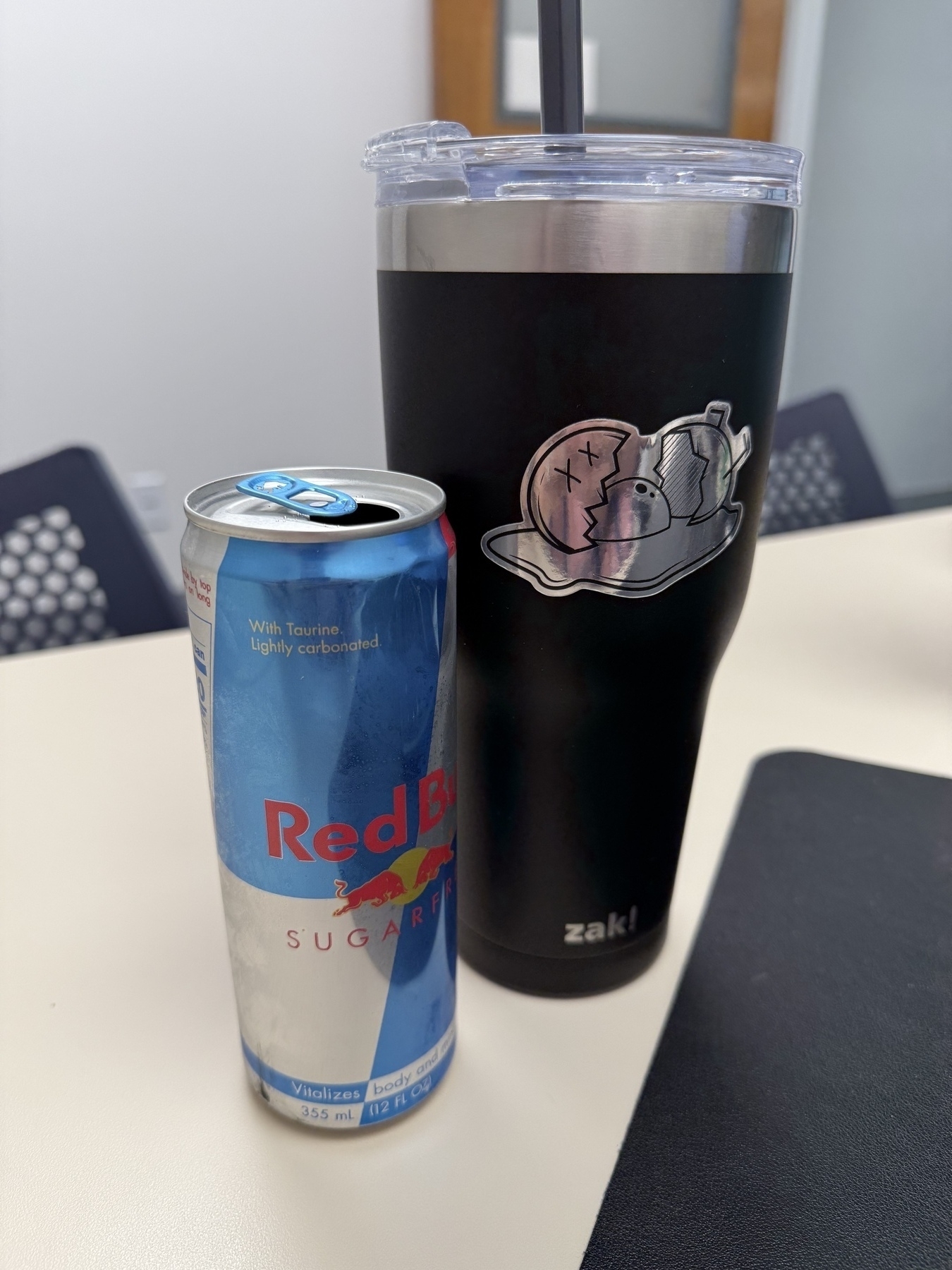 A can of Red Bull Sugarfree is placed next to a large Zak! tumbler with a cartoon sticker on a table.