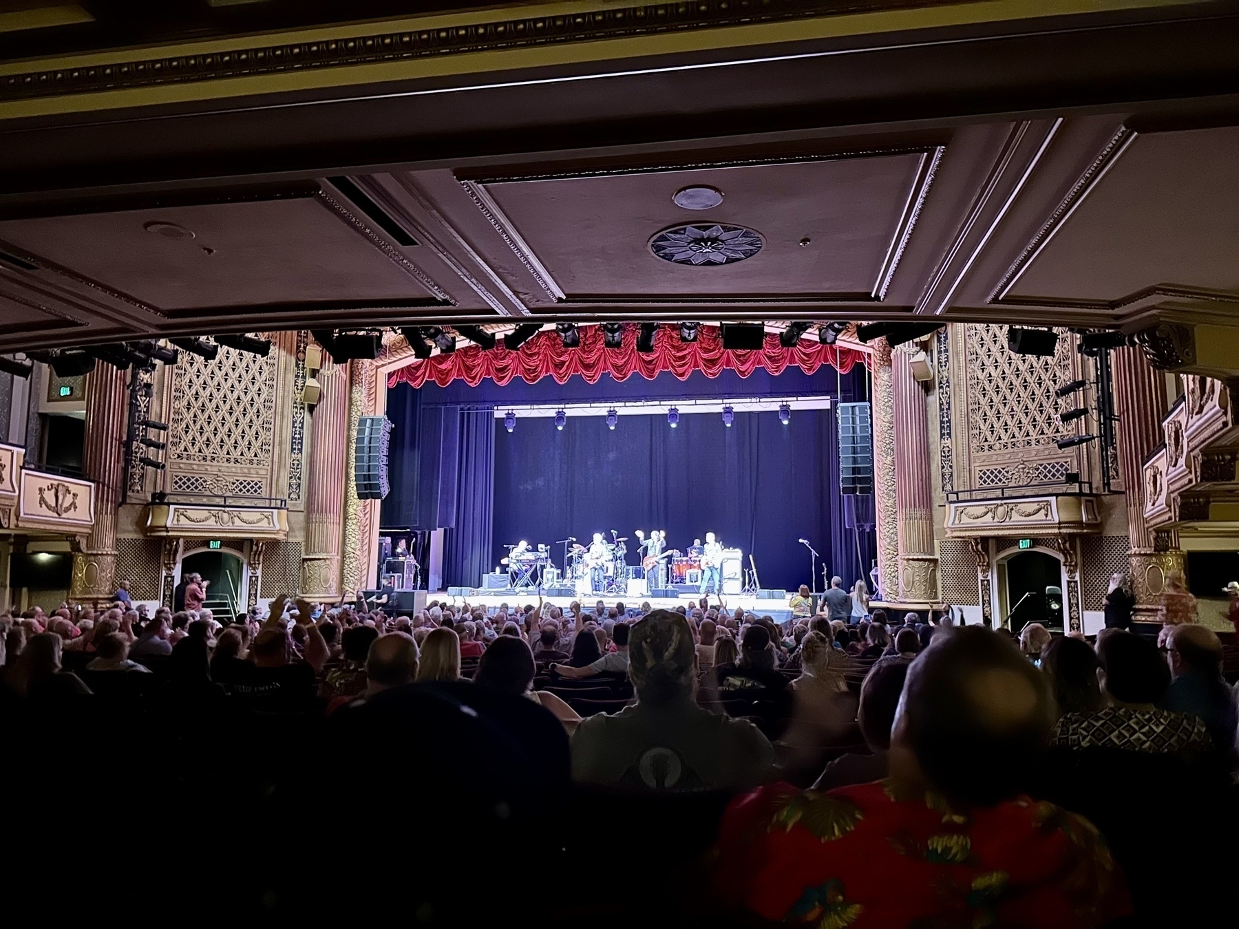 The image shows an indoor theater with an audience watching a live band performance on stage, featuring ornate architectural details and red curtains.