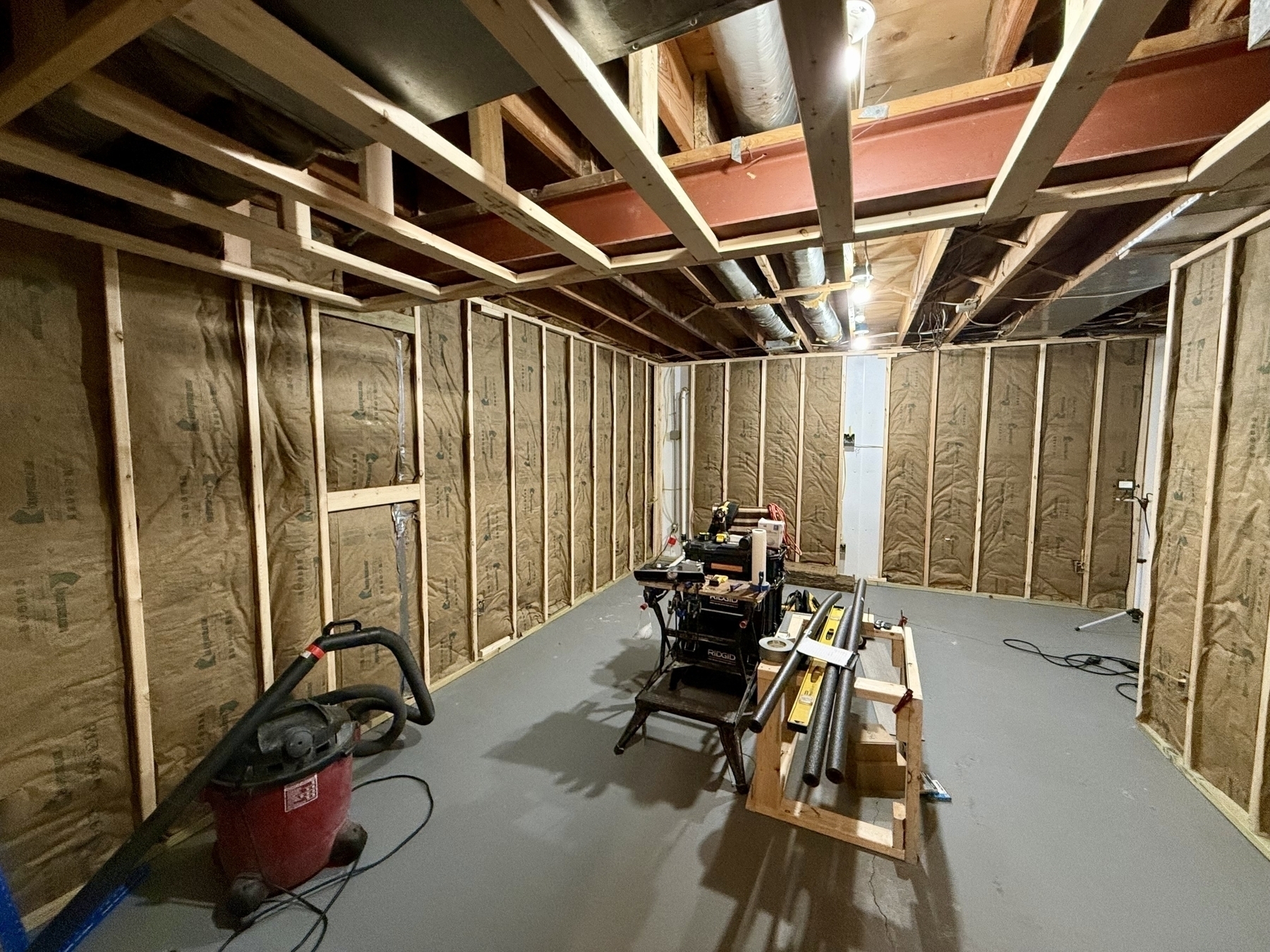 A partially finished basement features exposed insulation, a workbench with tools, and a shop vacuum on the concrete floor.