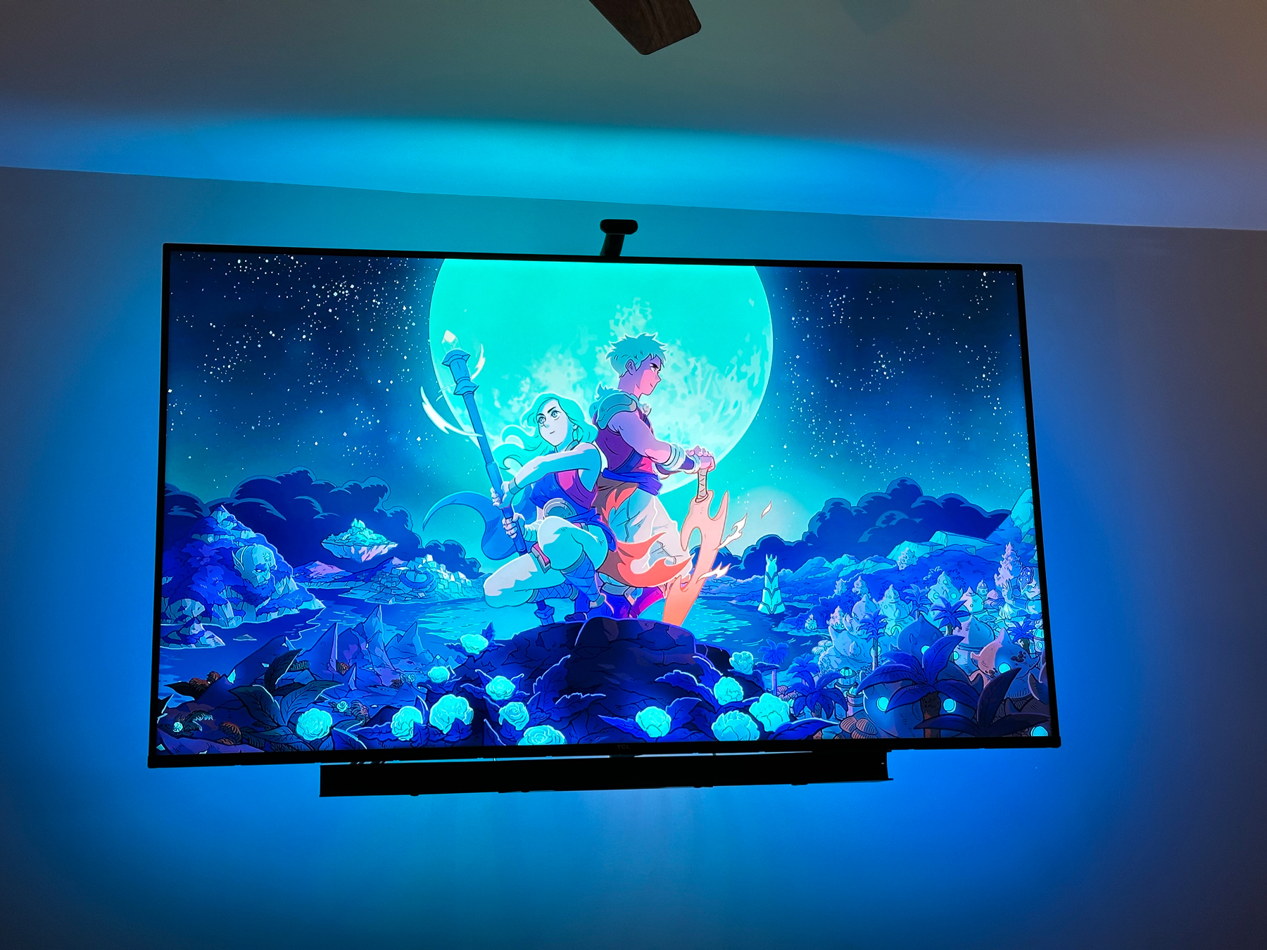 Animated fantasy artwork displayed on a screen, featuring a group of stylized characters in a vibrant blue and moonlit setting, surrounded by glowing flowers and a night sky filled with stars. Ambient blue lighting enhances the room’s atmosphere.
