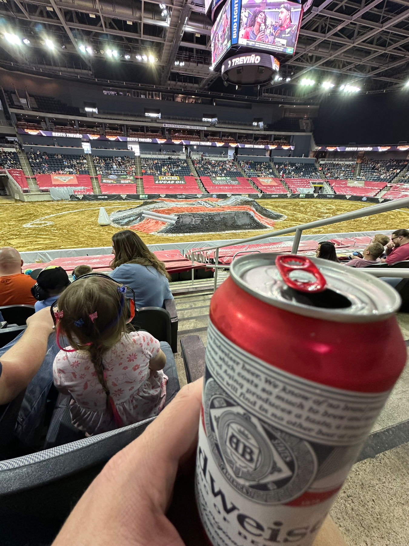 Person holding a canned beverage at a motorsport event in an arena, with dirt tracks prepared for competition. Spectators are seated, and a large overhead display shows event content.