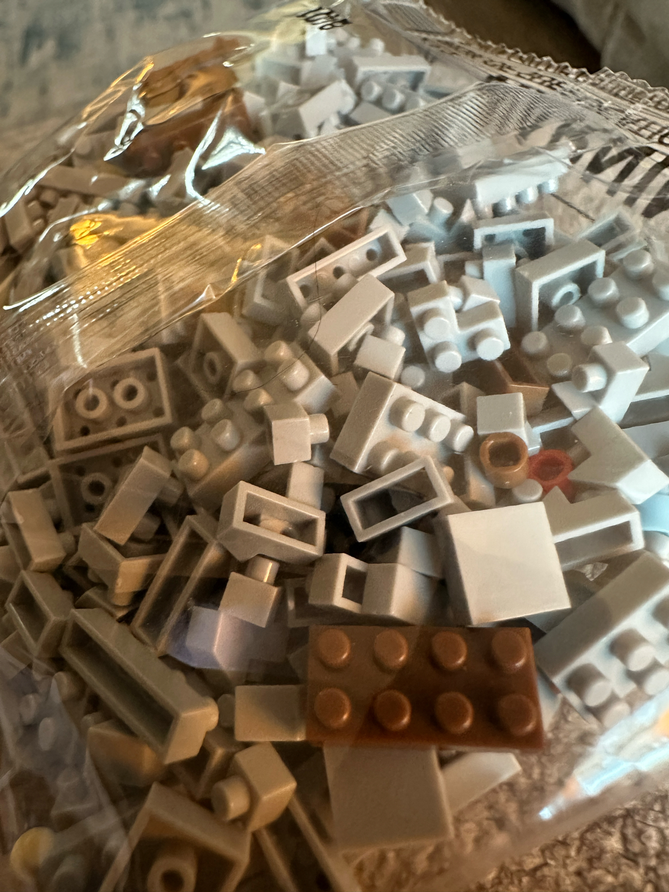 A bag of assorted LEGO bricks in various shapes and sizes, predominantly in different shades of gray and white, with a single brown brick visible.