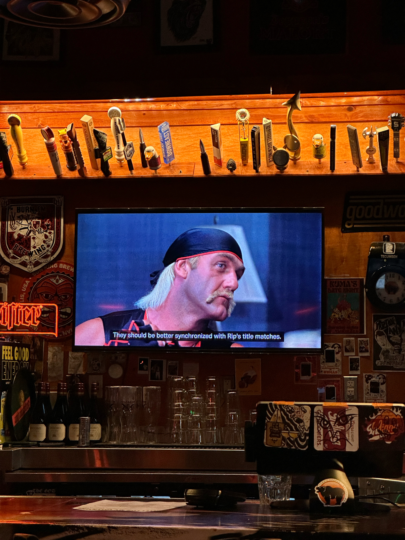 A television screen mounted in a bar showing a man with a bandana and mustache. Below the TV is a variety of draft beer taps and glasses on the counter. There’s a subtitle on the screen, and various posters and signs decorate the walls