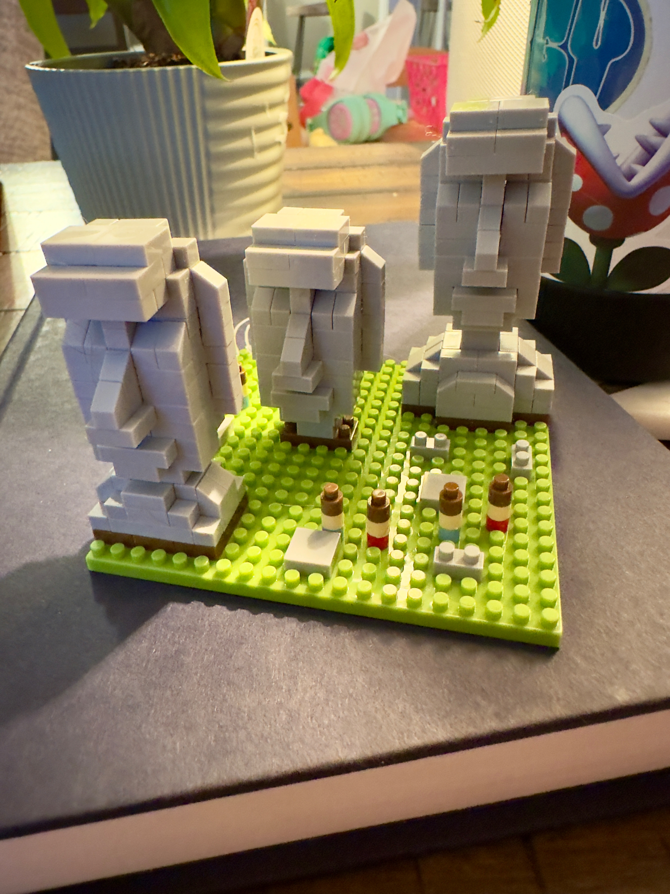 A LEGO model depicting three Moai statues (like those on Easter Island) on a green baseplate, surrounded by other miscellaneous LEGO pieces. In the background, there is a potted plant and other indistinct household items.