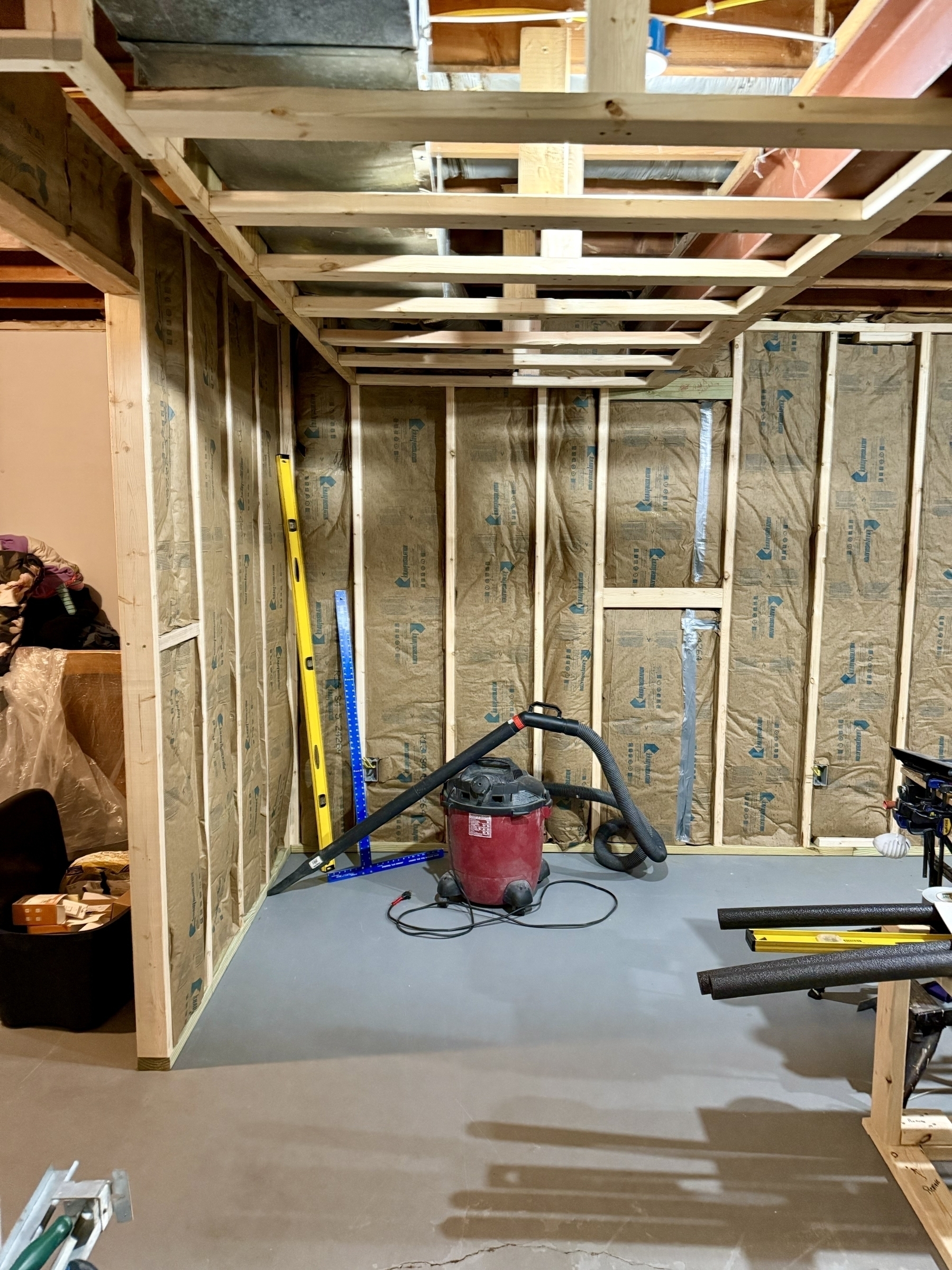 A construction area features exposed insulated walls, a shop vacuum, a level, and various tools scattered across a concrete floor.