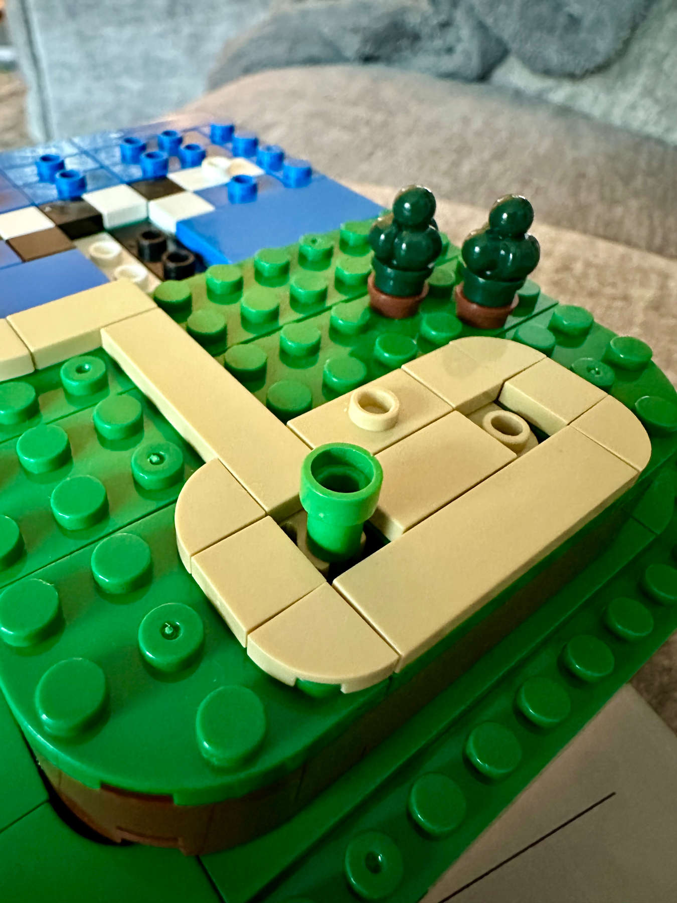 A close-up of a LEGO model featuring greenery, a beige pathway, and a blue and white water body. The focus is on the pathway and green LEGO plants.