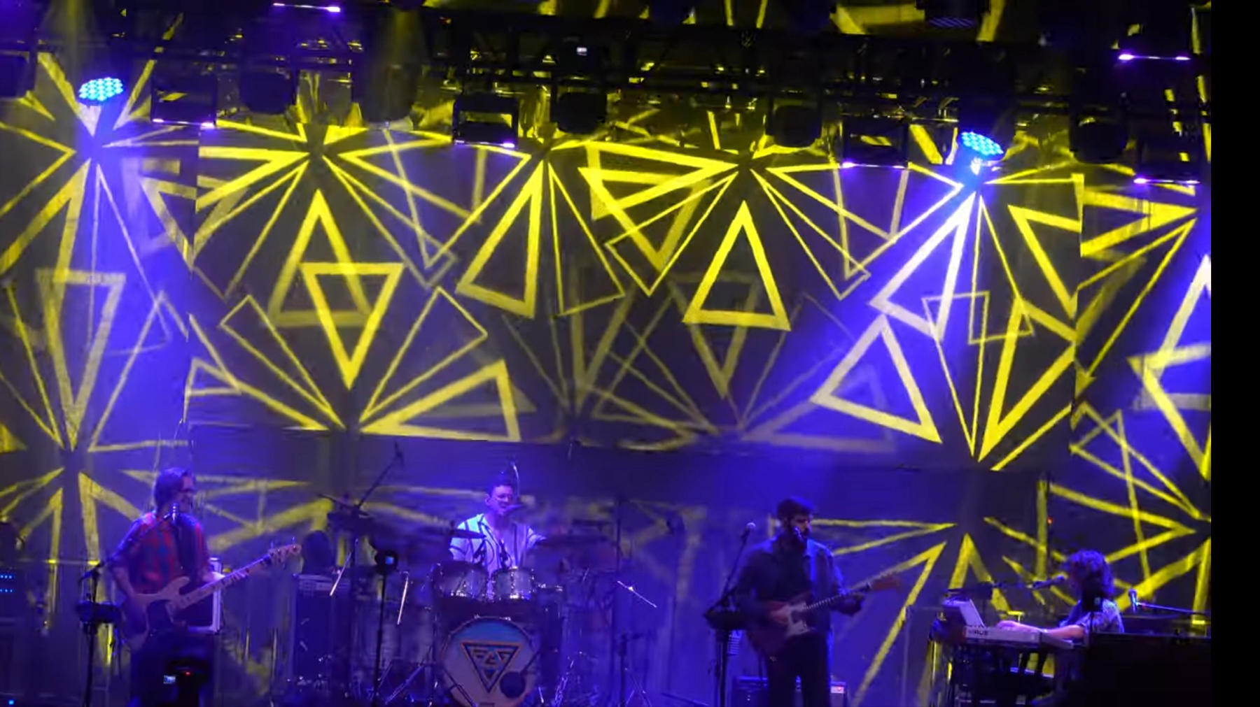 A band performing on stage with a dynamic lighting display featuring yellow geometric patterns in the background.
