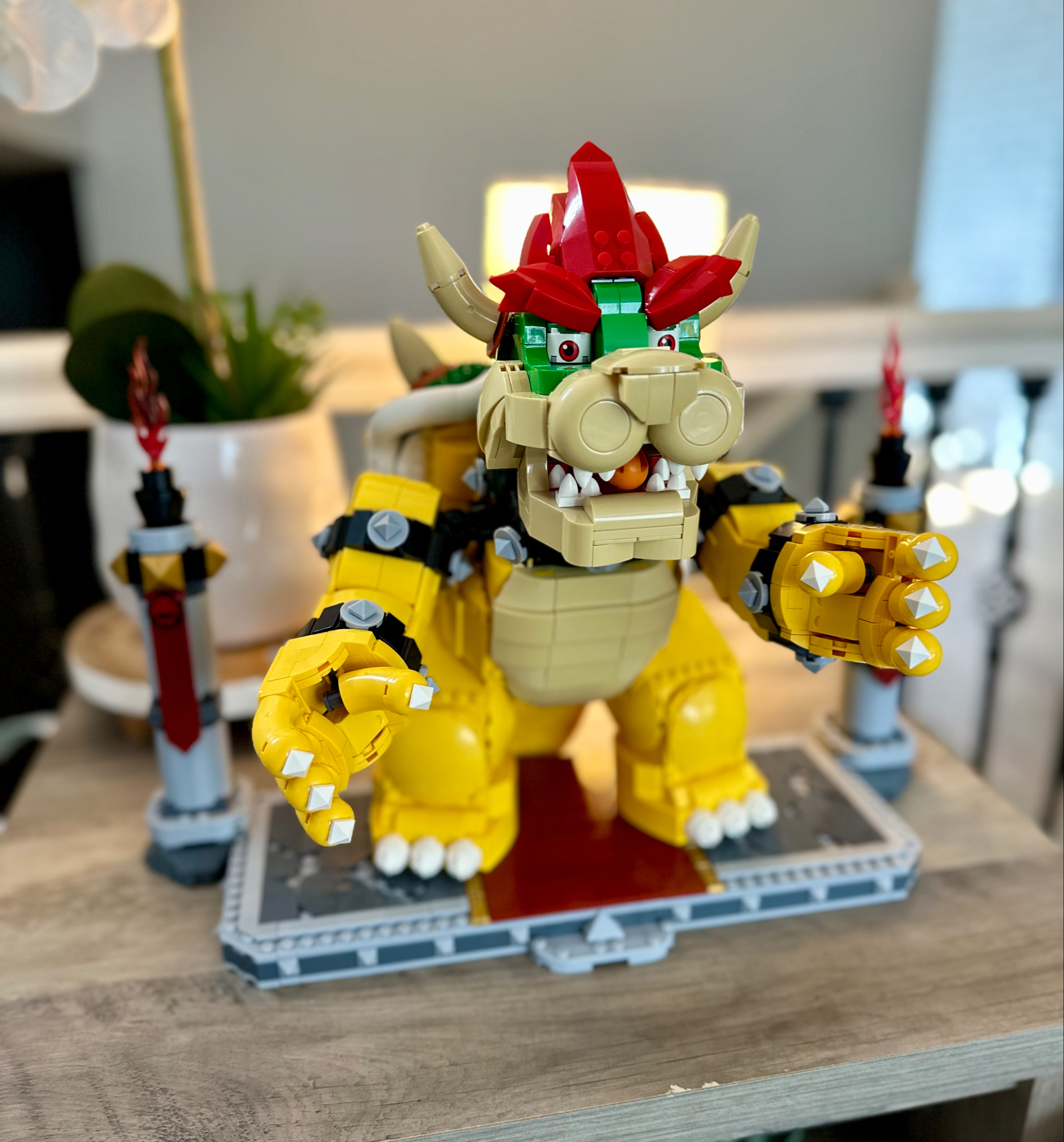 A LEGO model of a yellow and red dragon standing on a gray base, featuring green eyes, large claws, and an open mouth displaying white teeth. The backdrop includes a furnished room with indoor plants and a lamp.