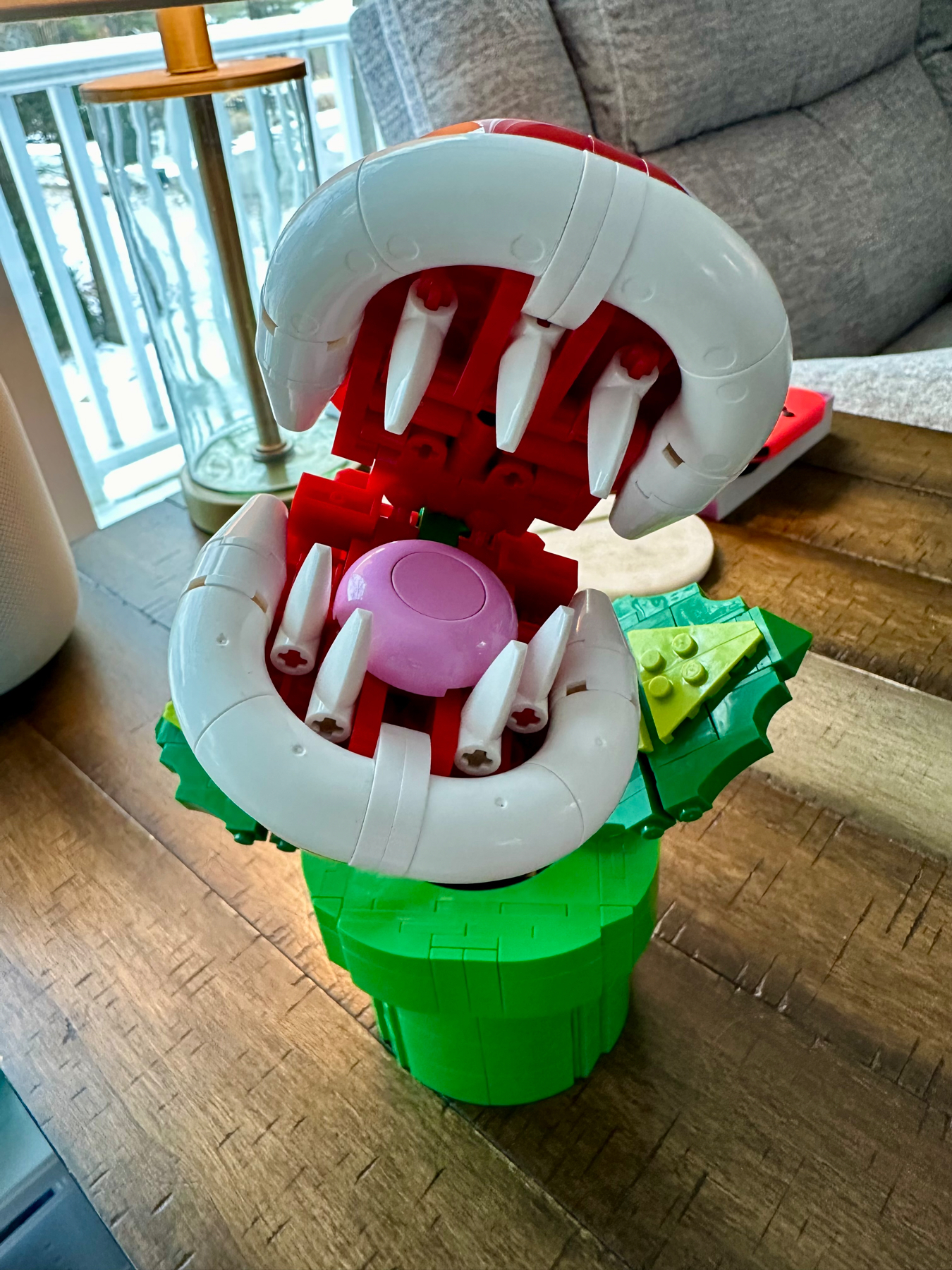 A colorful LEGO model of a flower in a green pot, featuring white petals with red details and a pink center, displayed indoors with a chair and balcony in the background.