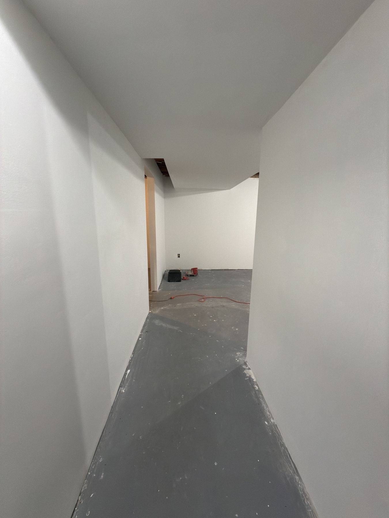 A minimalist interior space under construction features white walls, a gray floor, and an open ceiling, with construction equipment visible in the background.