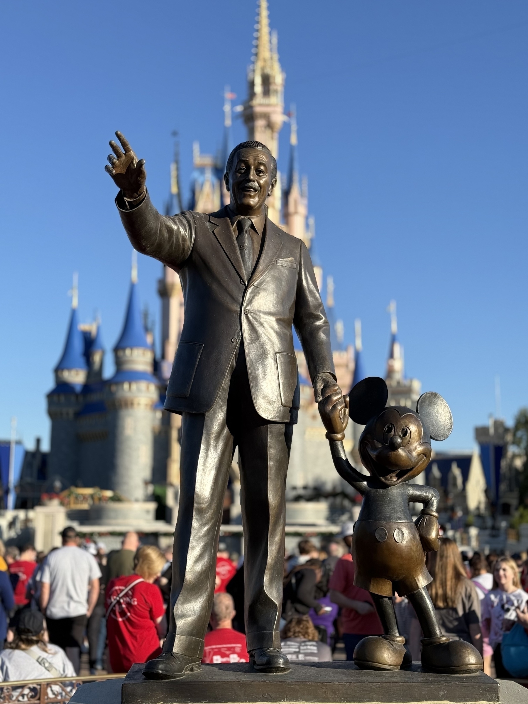 A statue of a man holding hands with a cartoon mouse stands in front of a castle, surrounded by people.