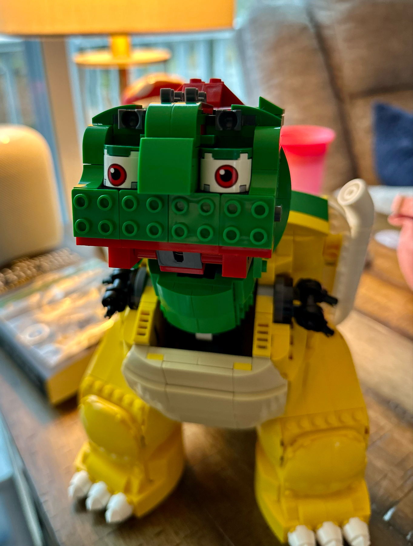 A close-up of a LEGO model dinosaur displaying its head and upper body. The dinosaur is predominantly green with touches of red and white, and has posable arms with black claws. The background is blurred with indistinct household items.