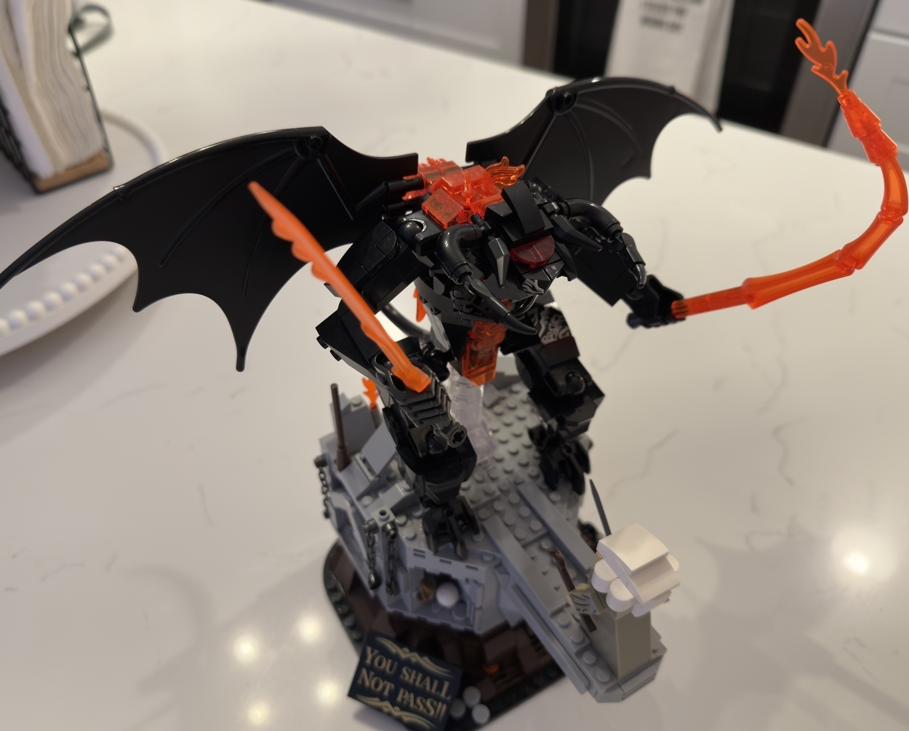 A LEGO model of a winged creature holding fiery weapons stands atop a rocky structure with a small plaque.